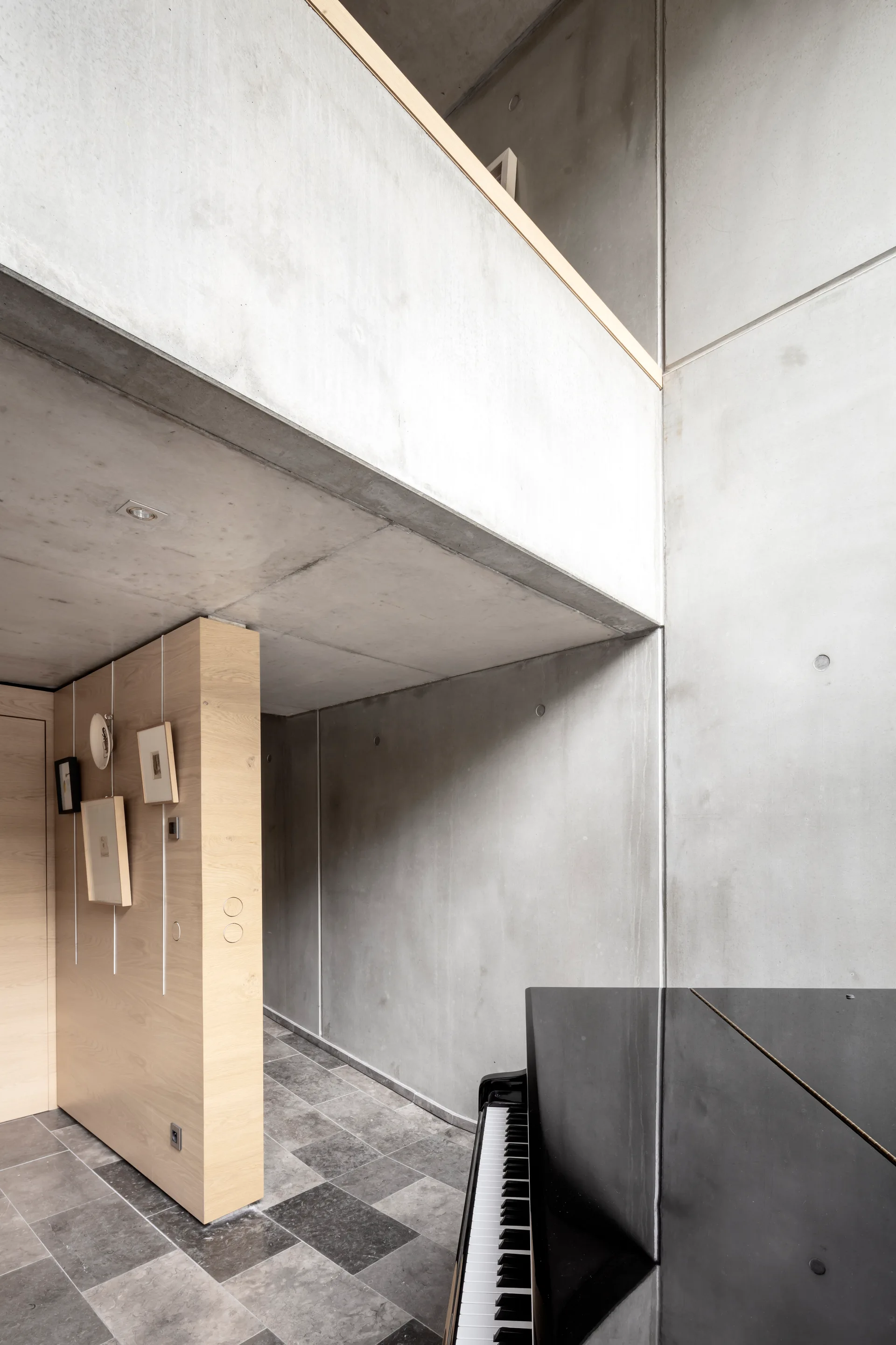 A modern interior featuring raw concrete walls, a light wood partition hanging small pictures, a grey stone tile floor, and a black piano in the foreground.