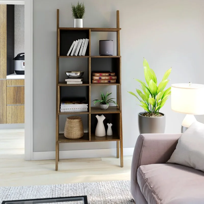A wooden ladder bookshelf with two-tone compartments stands against a gray wall next to a potted plant, sofa, and lamp.