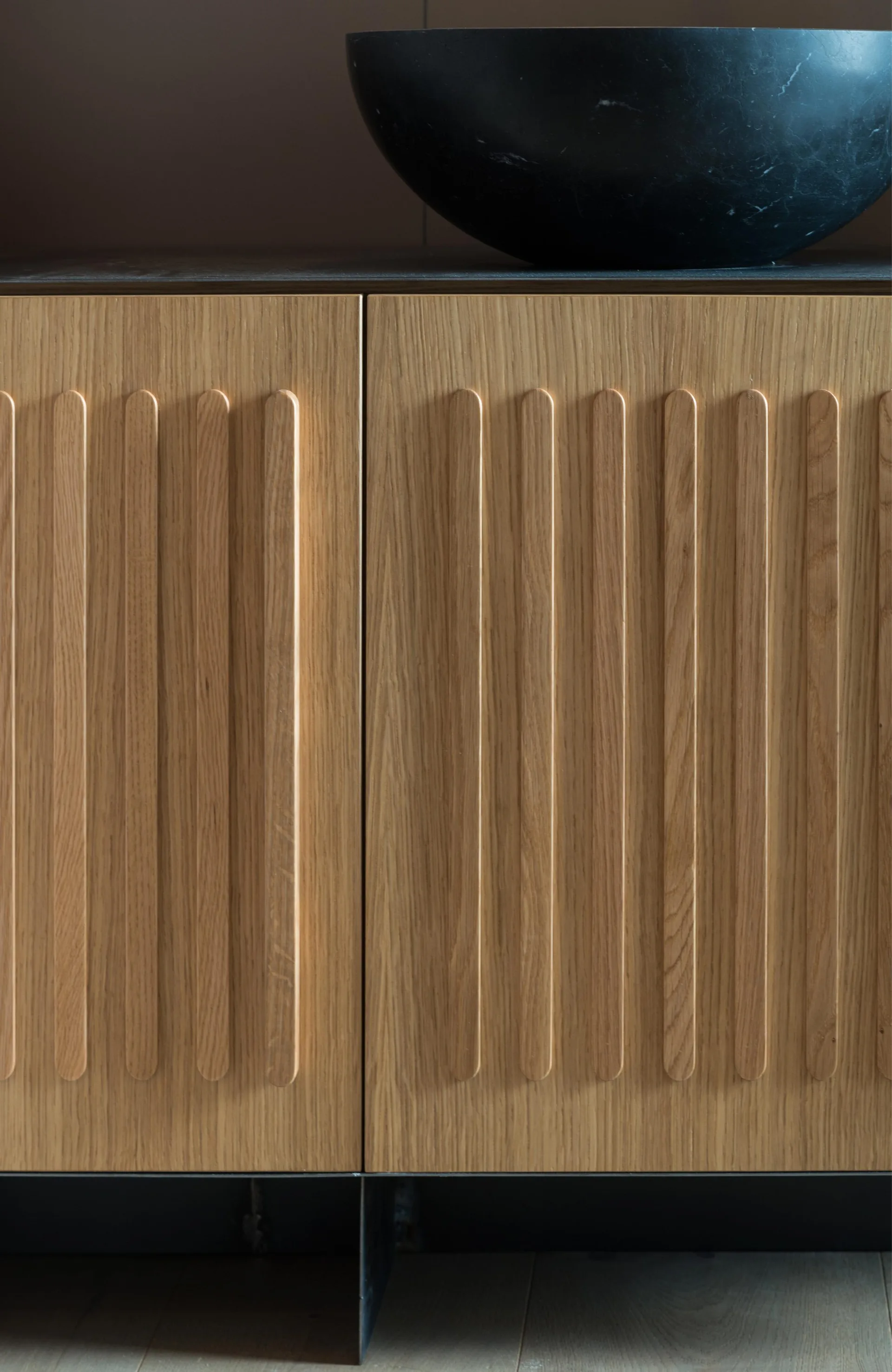 Close-up of light oak cabinet doors with rounded vertical fluting details beneath a black stone vessel sink.
