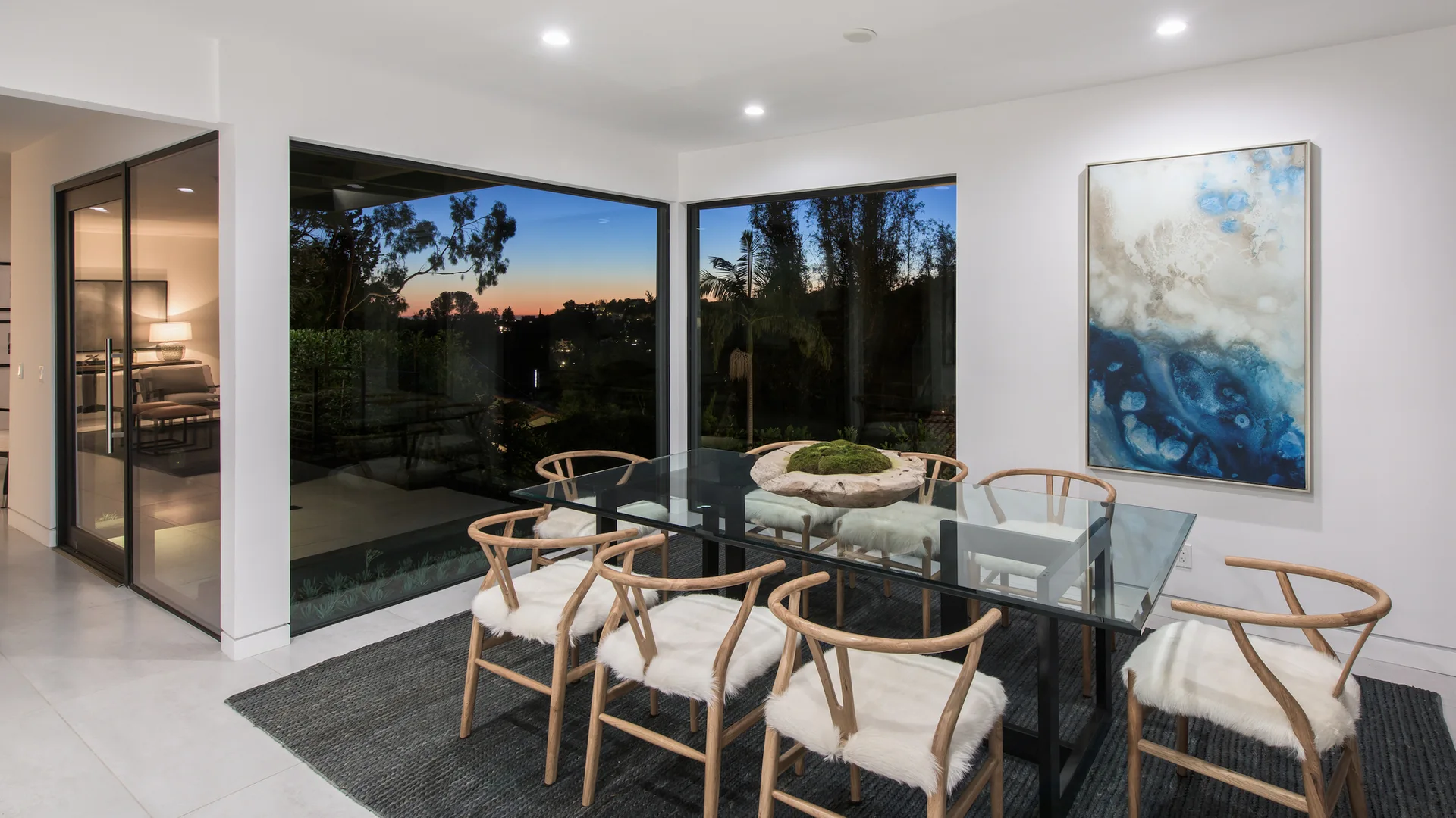 A modern dining room features a glass-top table, light wood chairs with white faux fur pads, and large corner windows displaying a twilight view.