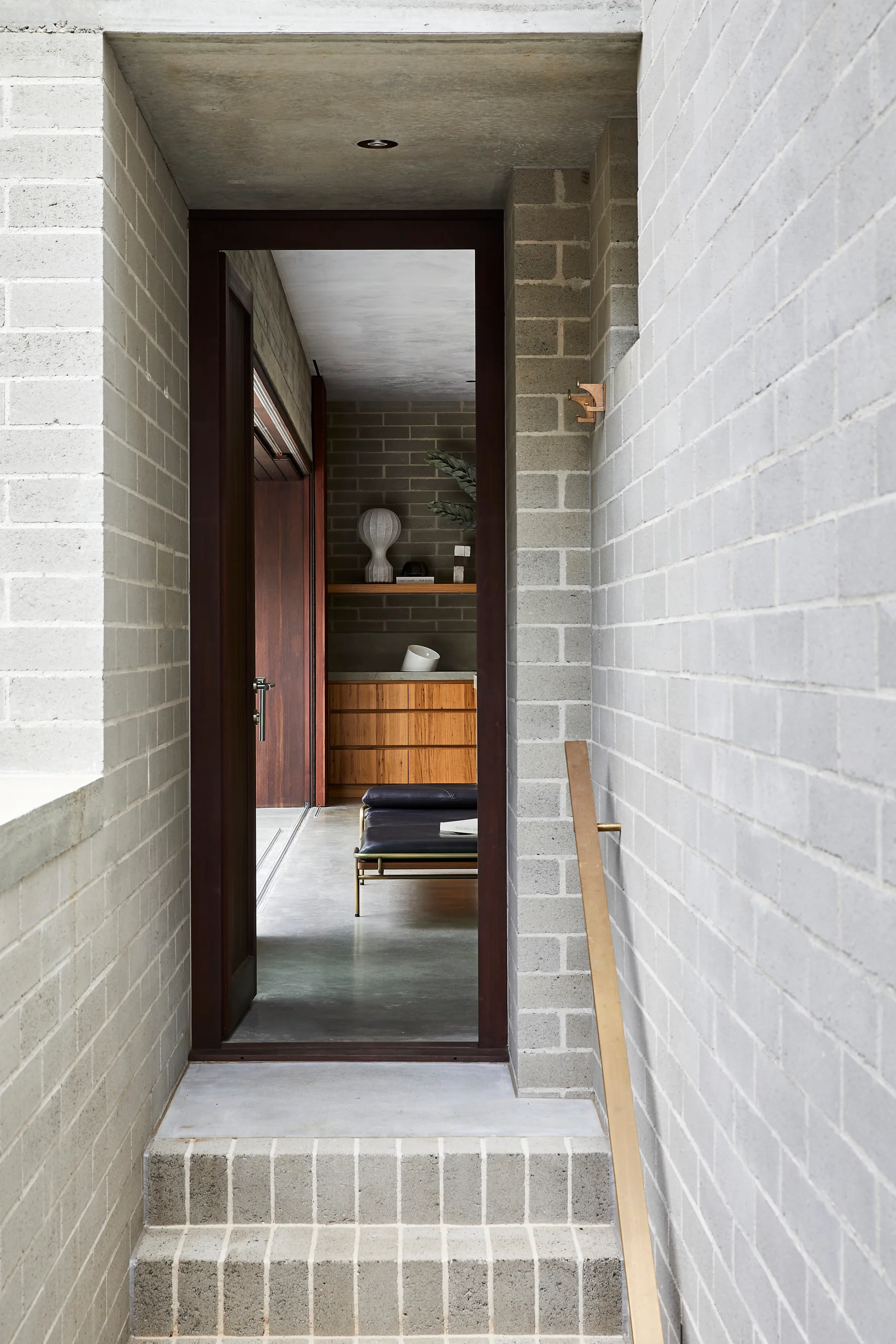 A transitional hallway features light grey brick walls, concrete steps, and a wooden handrail leading to a doorway that reveals a wood-toned interior.