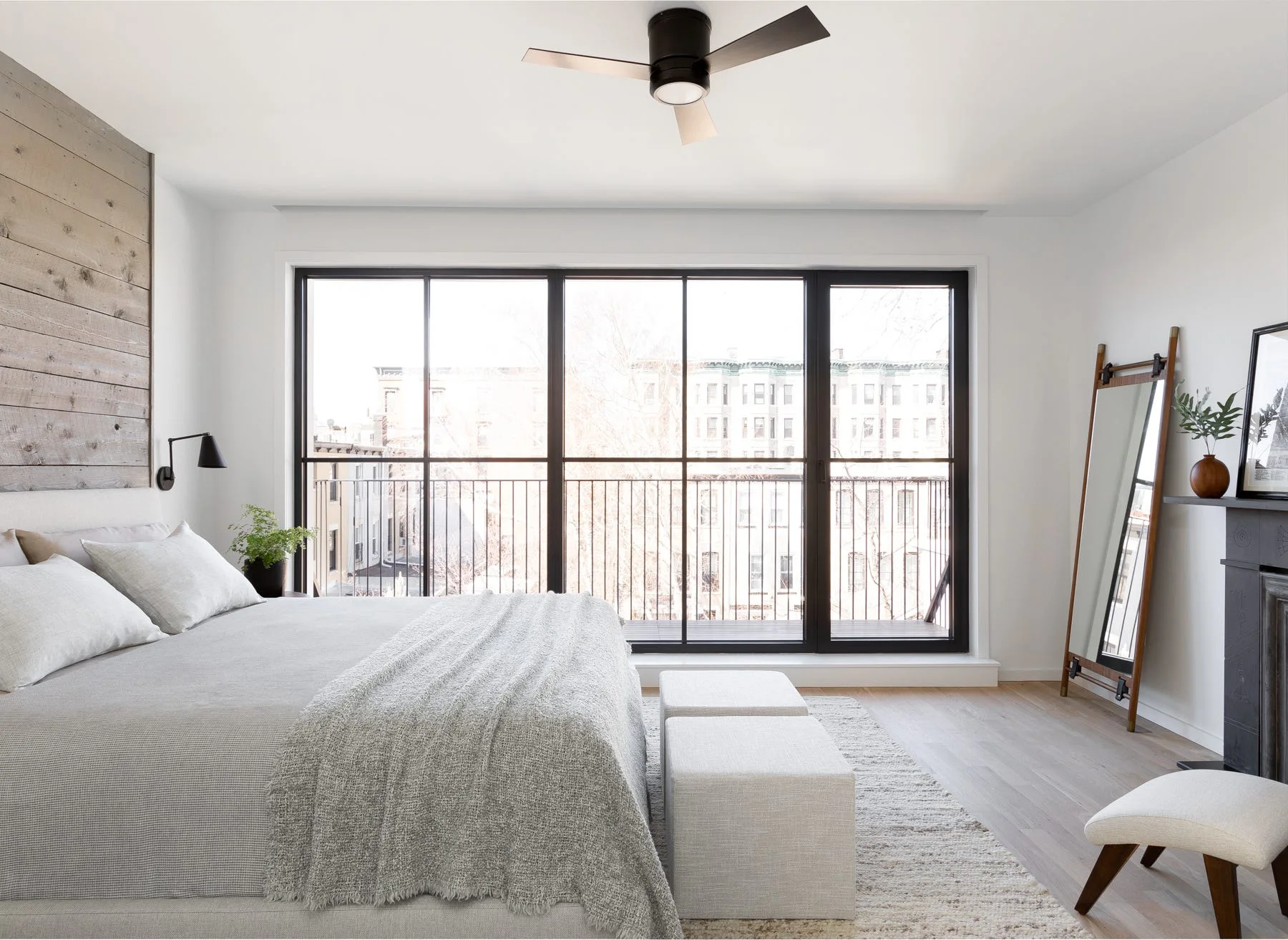 A bright bedroom with grey bedding, a weathered wood plank accent wall, large black-framed balcony windows, and a leaning floor mirror.