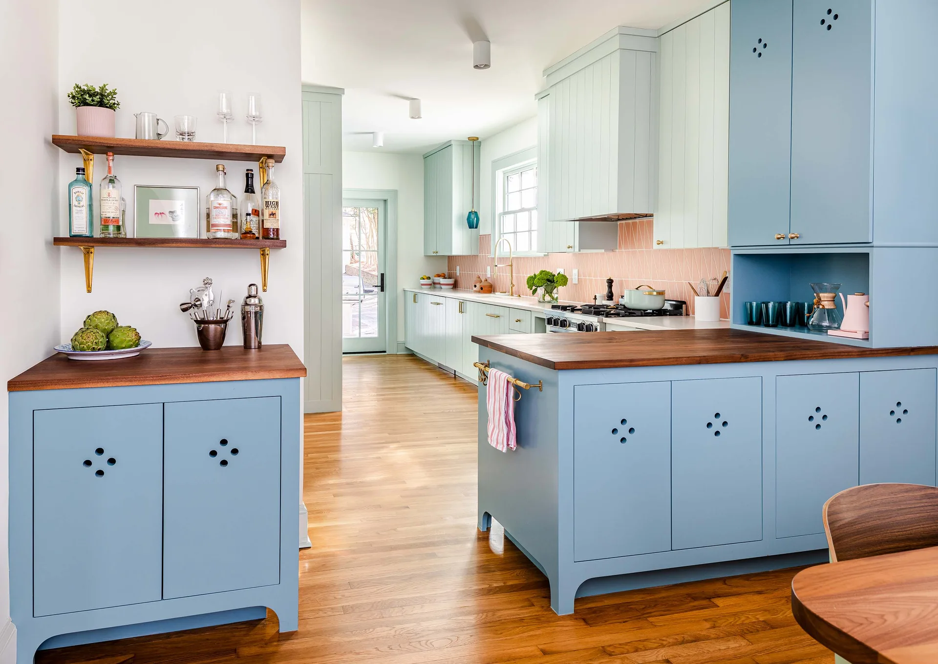 A bright kitchen features blue and pale green cabinets with warm wood countertops, a pink tile backsplash, and light wood flooring.