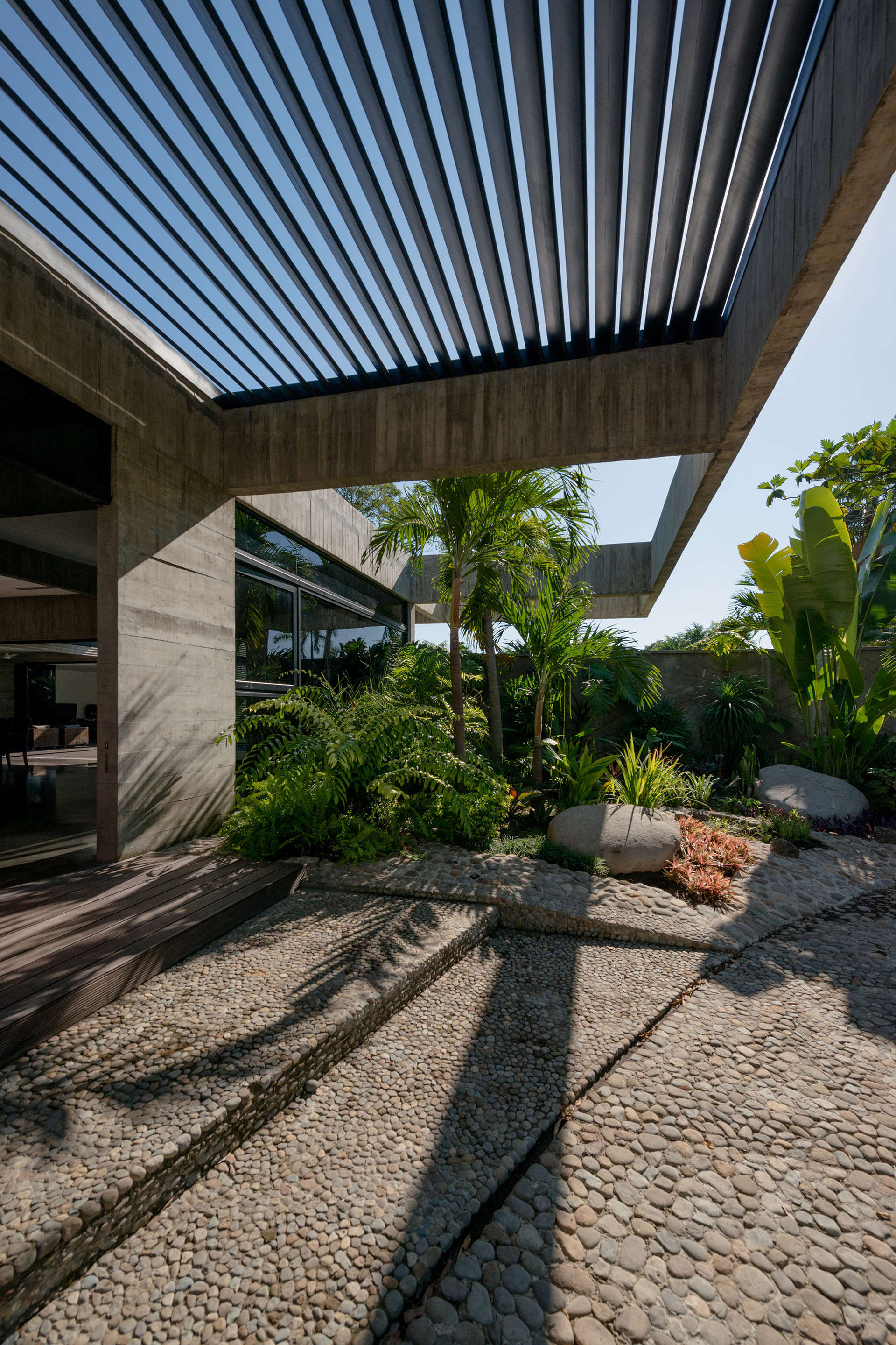 A modern concrete pergola with a dark louvred roof frames a pebble-paved patio steps away from a lush tropical garden.