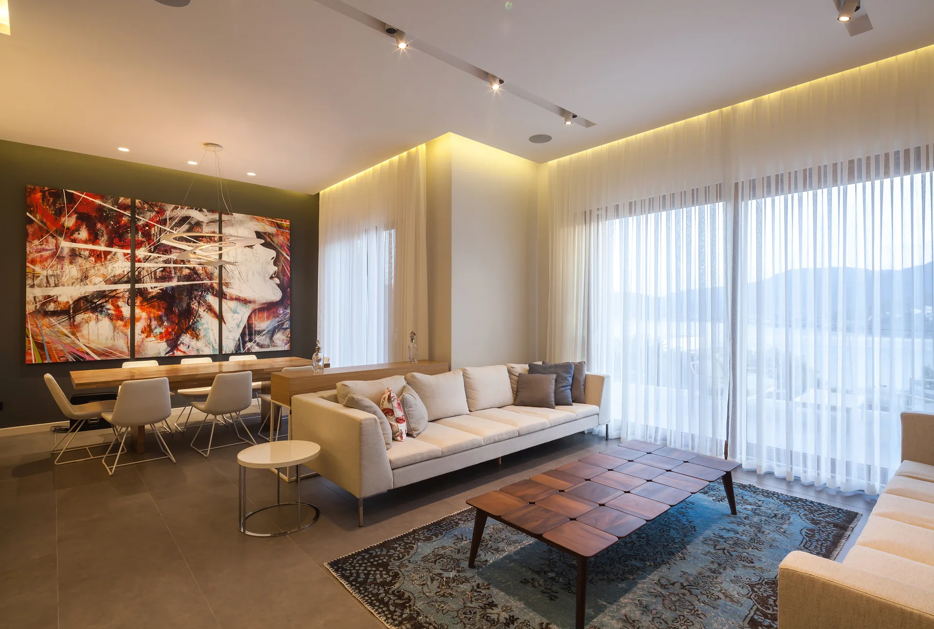 A modern open-plan living room features a beige sofa, a paneled wood coffee table on a patterned blue rug, and a dining area under multi-panel wall art.