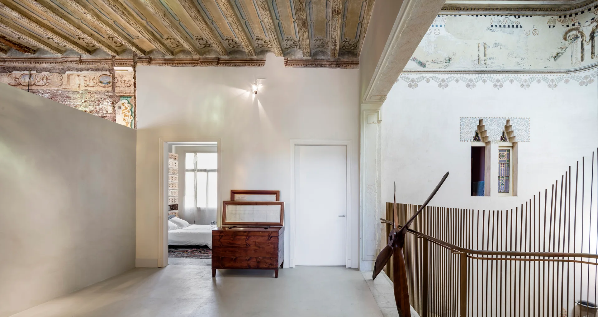 A modern hallway landing featuring a historic painted wood ceiling, a simple wooden chest, a slatted metal railing, and a view into a bedroom.