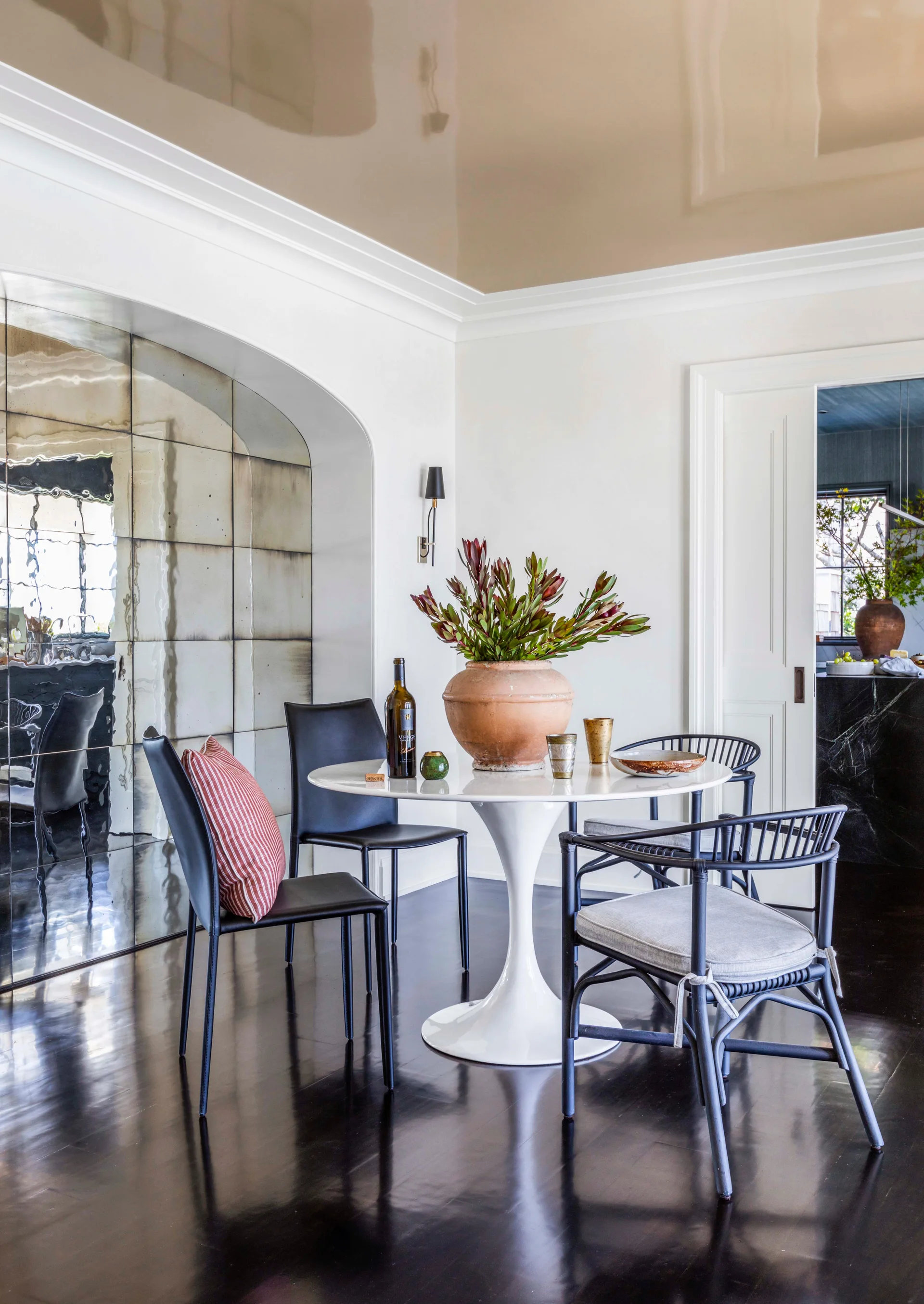 A white pedestal table and mixed black chairs sit on a dark glossy floor next to an arched wall lined with antique mirror tiles.