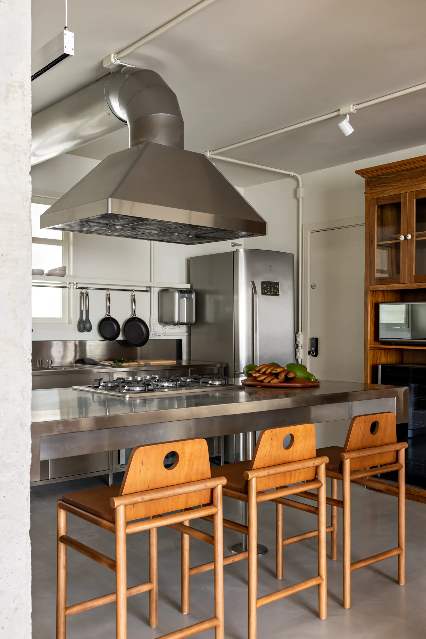 Three wooden bar stools sit at a stainless steel kitchen island with a large industrial vent hood overhead.