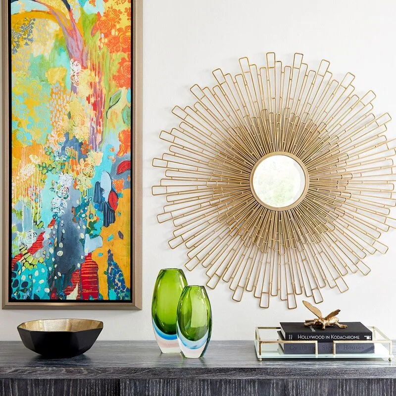 A dark wood console table displays green glass vases and books beneath a large gold sunburst mirror and colorful abstract painting on a white wall.