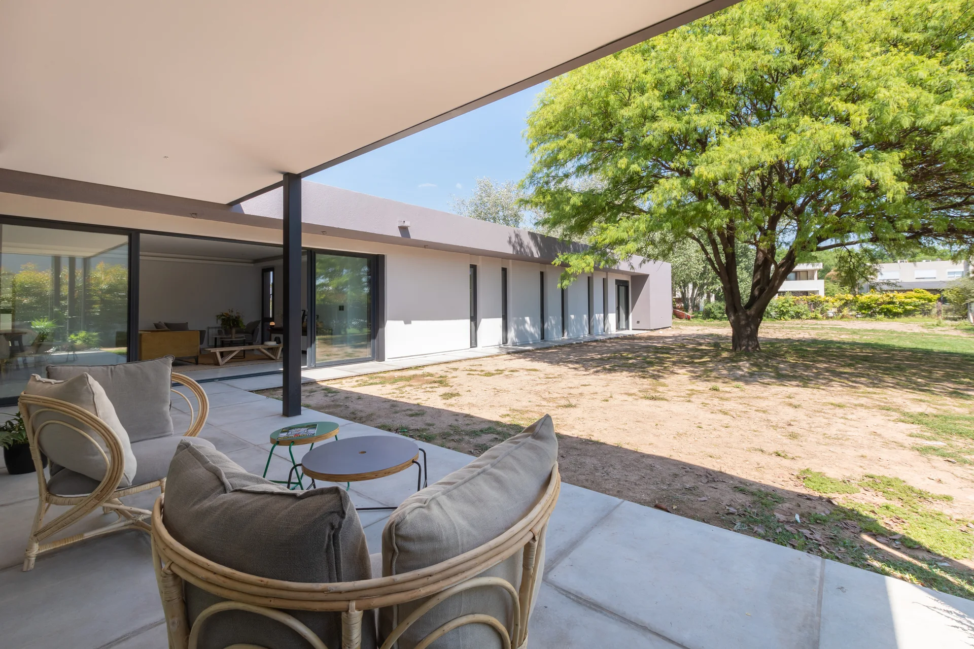A modern covered concrete patio features two curved rattan armchairs with beige cushions overlooking a large sunny lawn with a mature green tree.