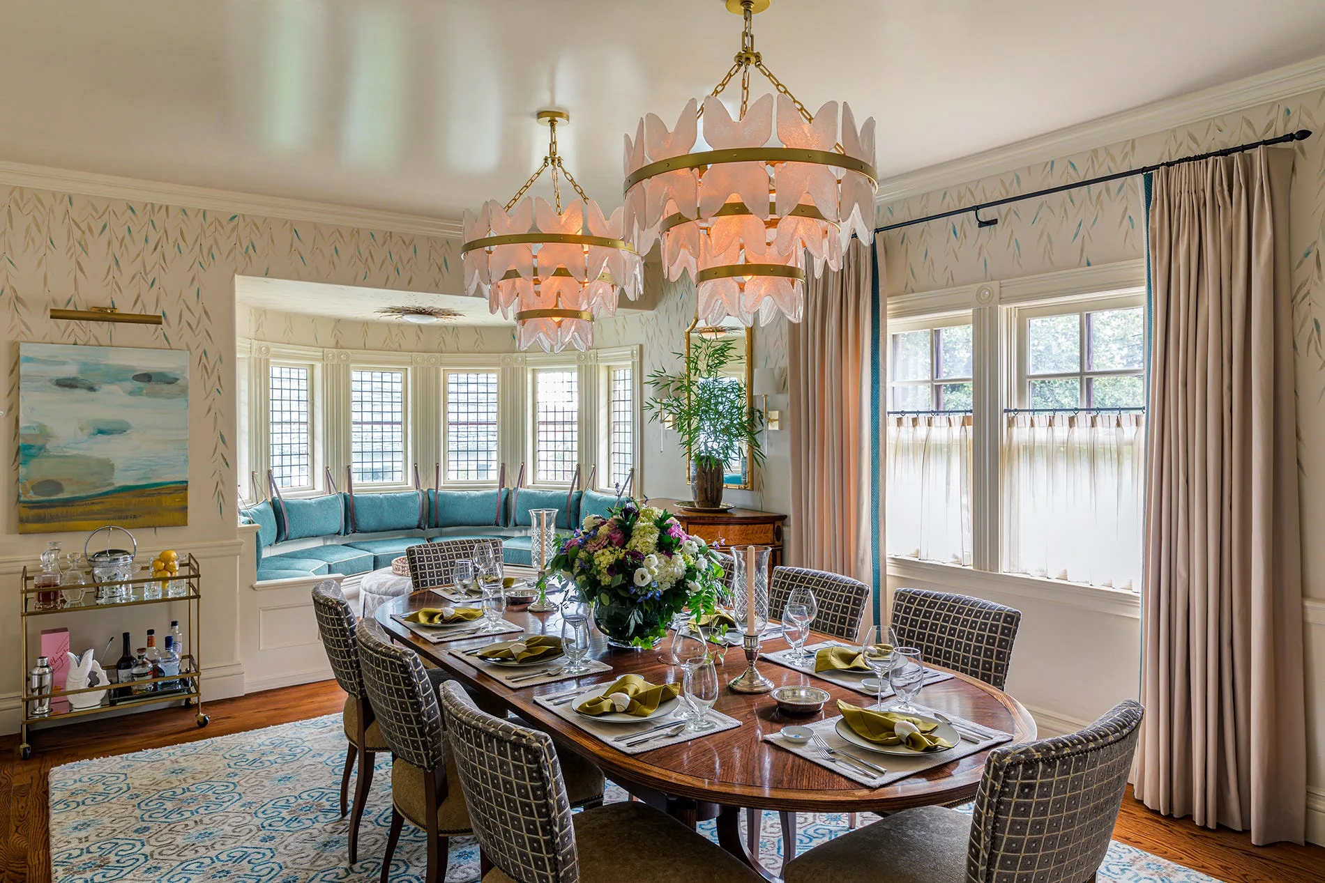 An oval wood dining table is surrounded by patterned chairs beneath two gold petal chandeliers, with a teal window seat in the background.