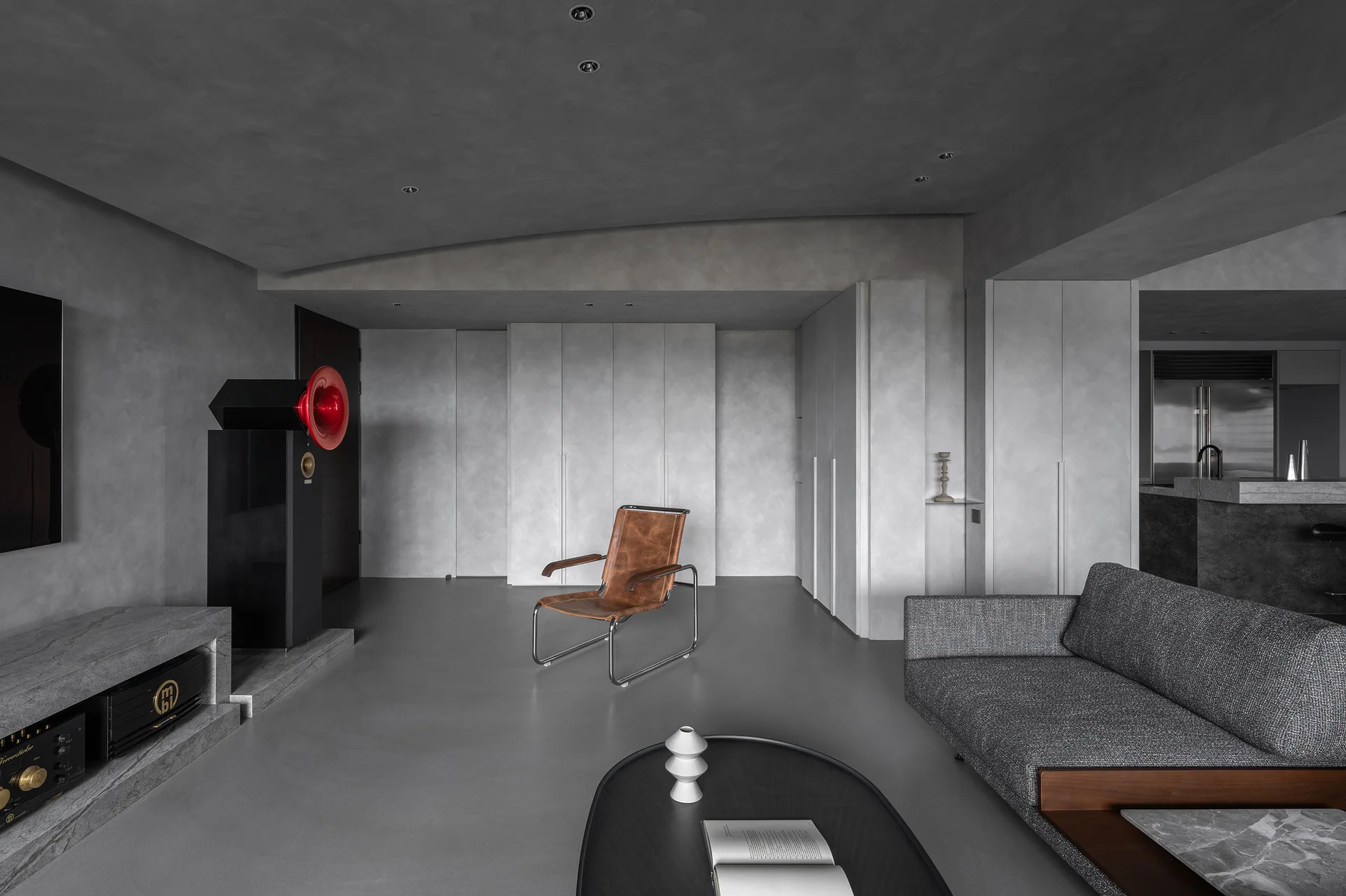 A minimalist living room features grey plaster walls and flooring, a dark grey fabric sofa, a tan leather armchair, and a prominent black speaker.