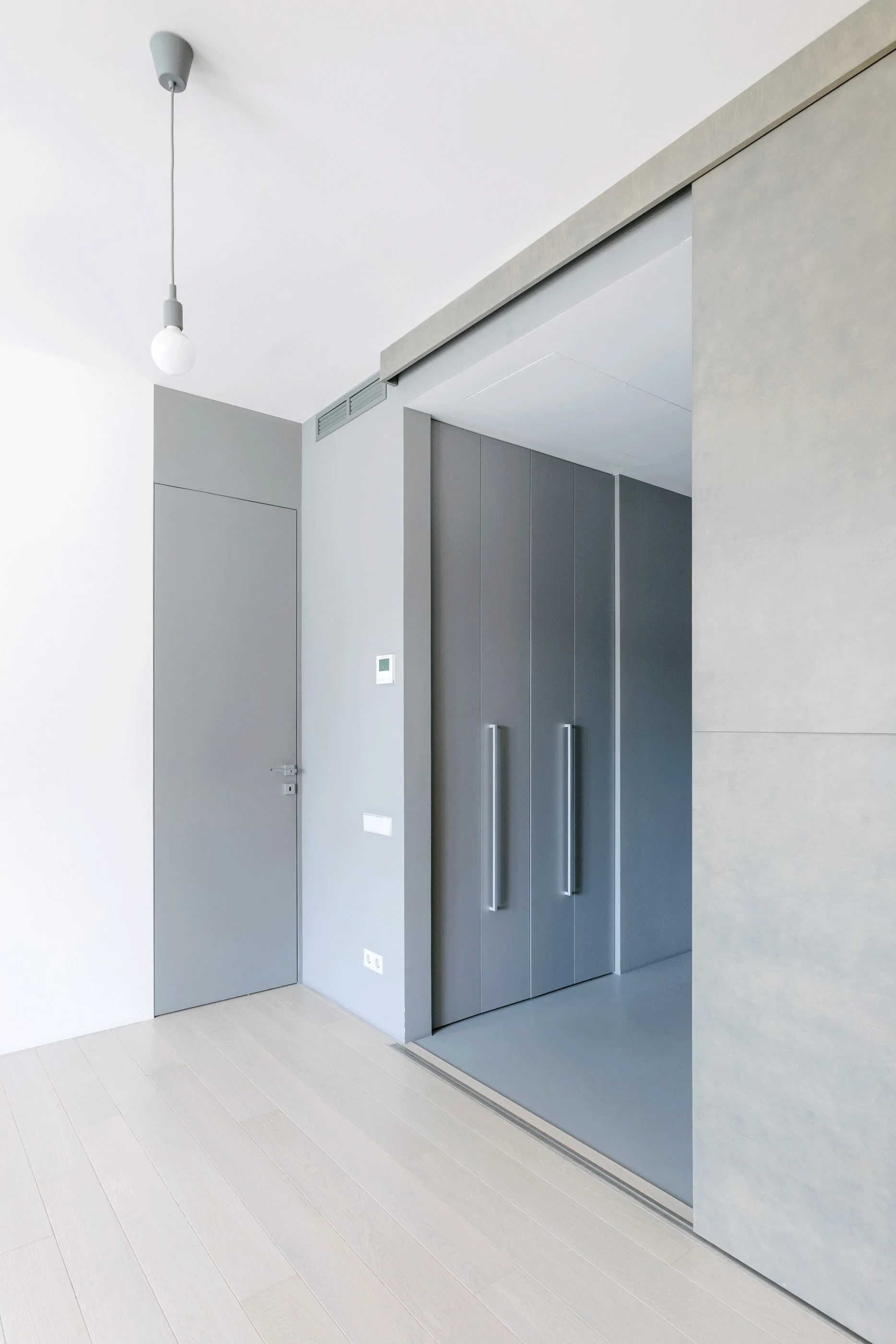 A minimalist gray hallway features a flush door, a large sliding partition, and a tall built-in closet with vertical handles over light wood flooring.