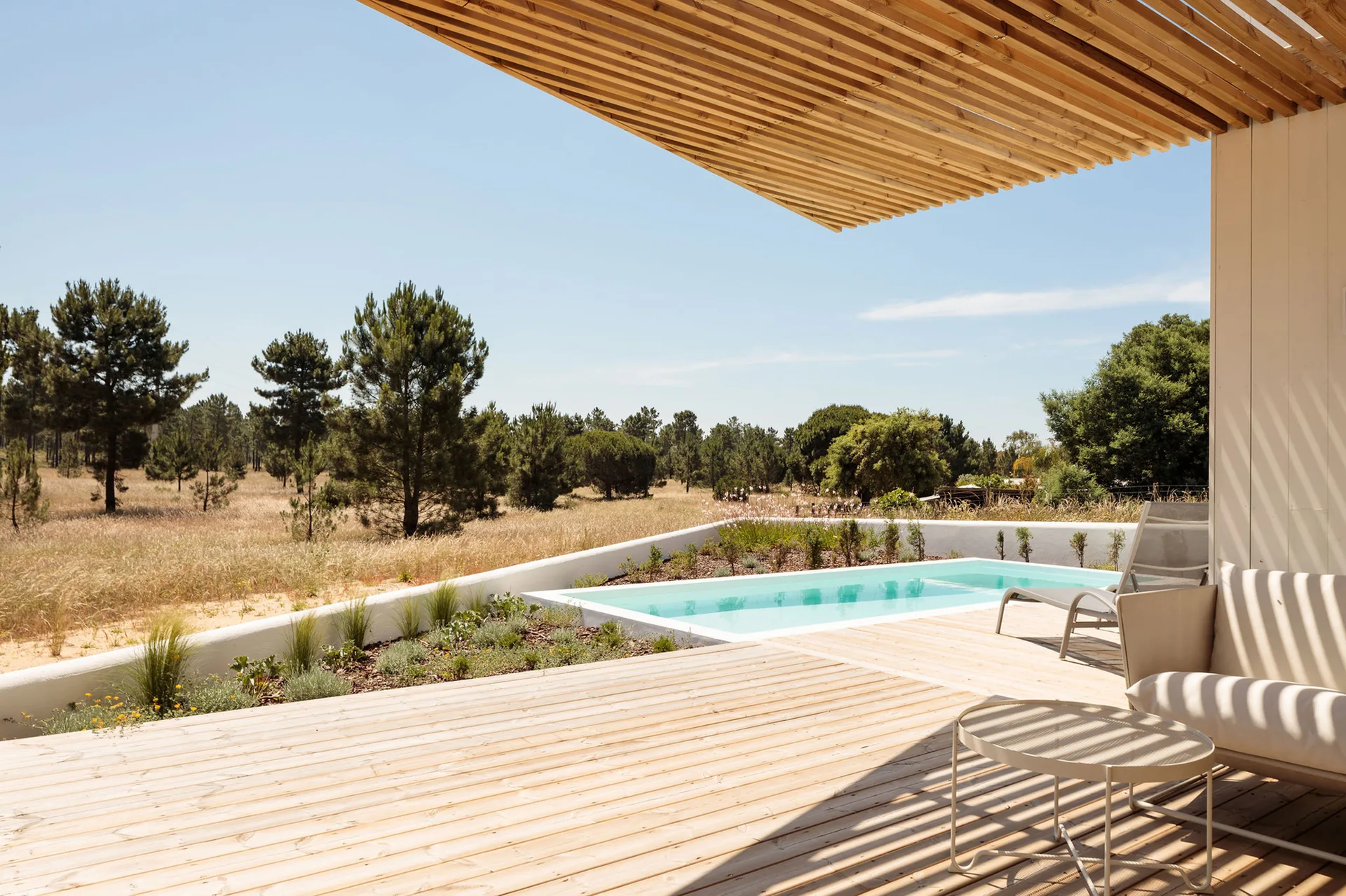 A sunny outdoor pool deck featuring a rectangular swimming pool, metal lounge chairs, a low white wall, and a slatted wood overhang casting shadows.