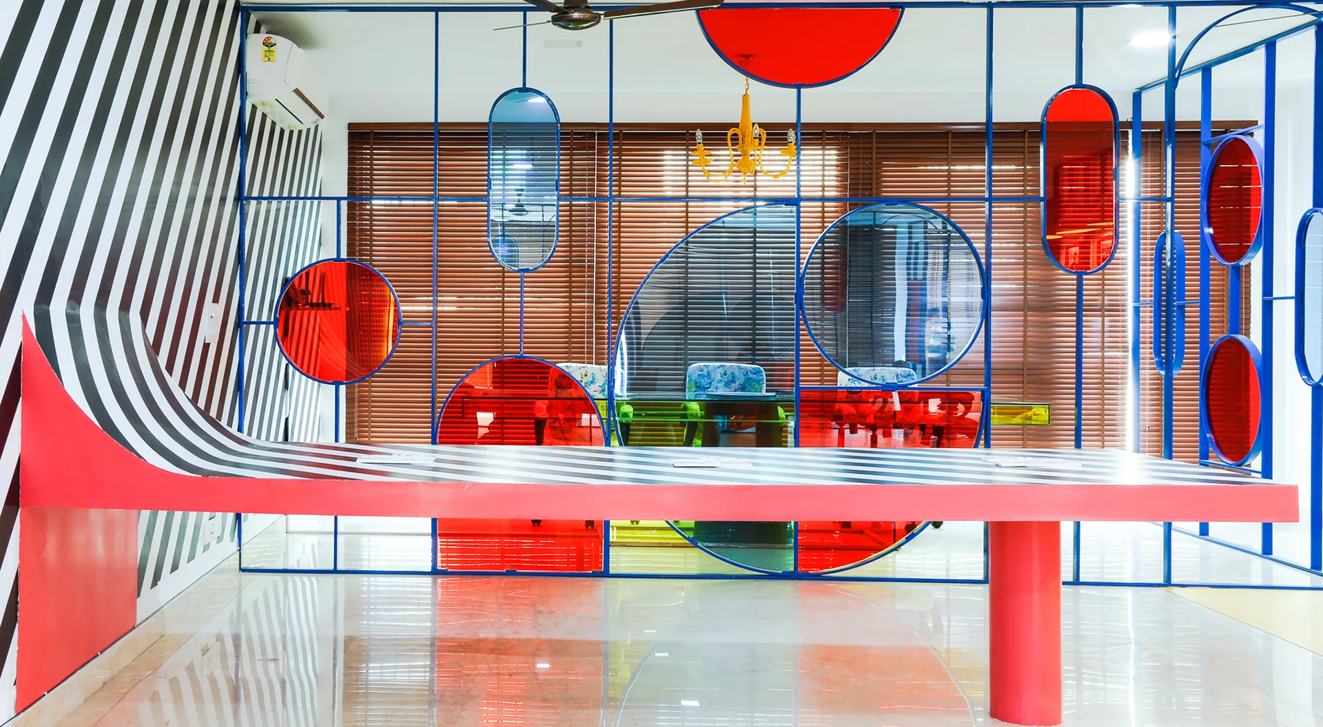A dining room features a long table with black and white stripes and a room partition made of blue metal frames with red and blue glass inserts.