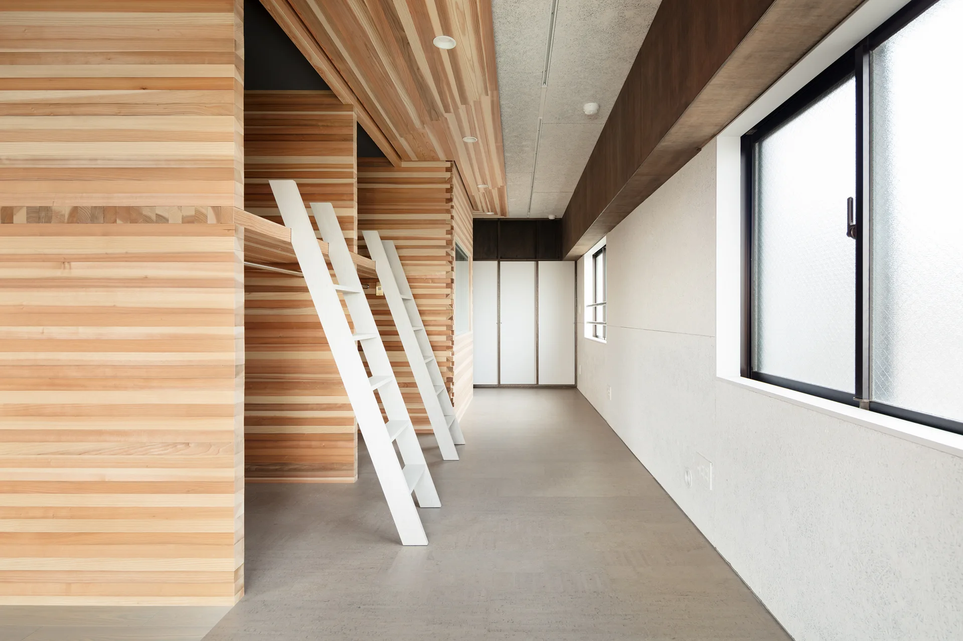 Two integrated wooden bunk bed structures with horizontal slats and white ladders stand in a minimalist room with gray floors and white walls.