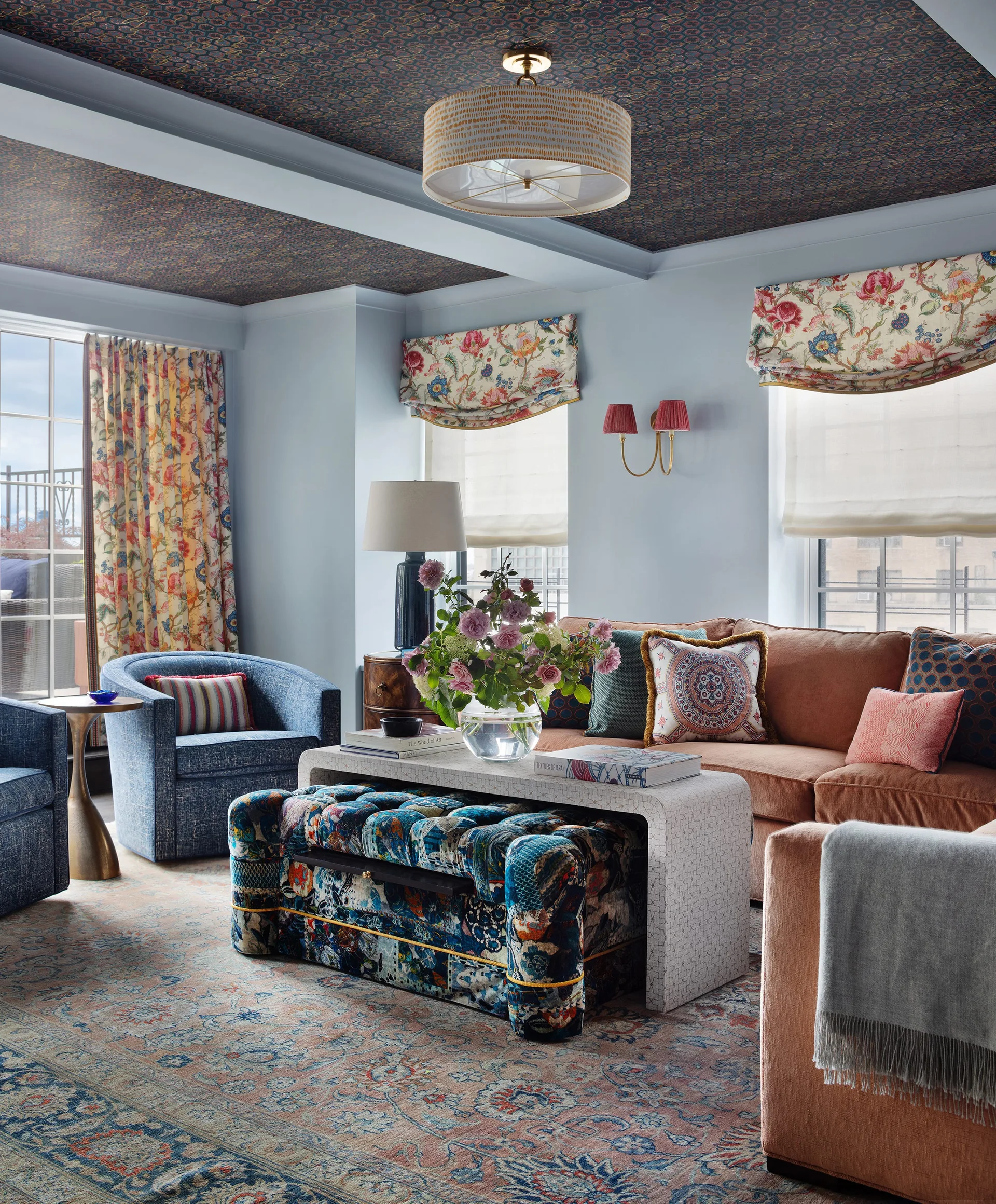 A living room with pale blue walls, a floral patterned ceiling, a tan velvet sofa, and blue armchairs around a white coffee table.