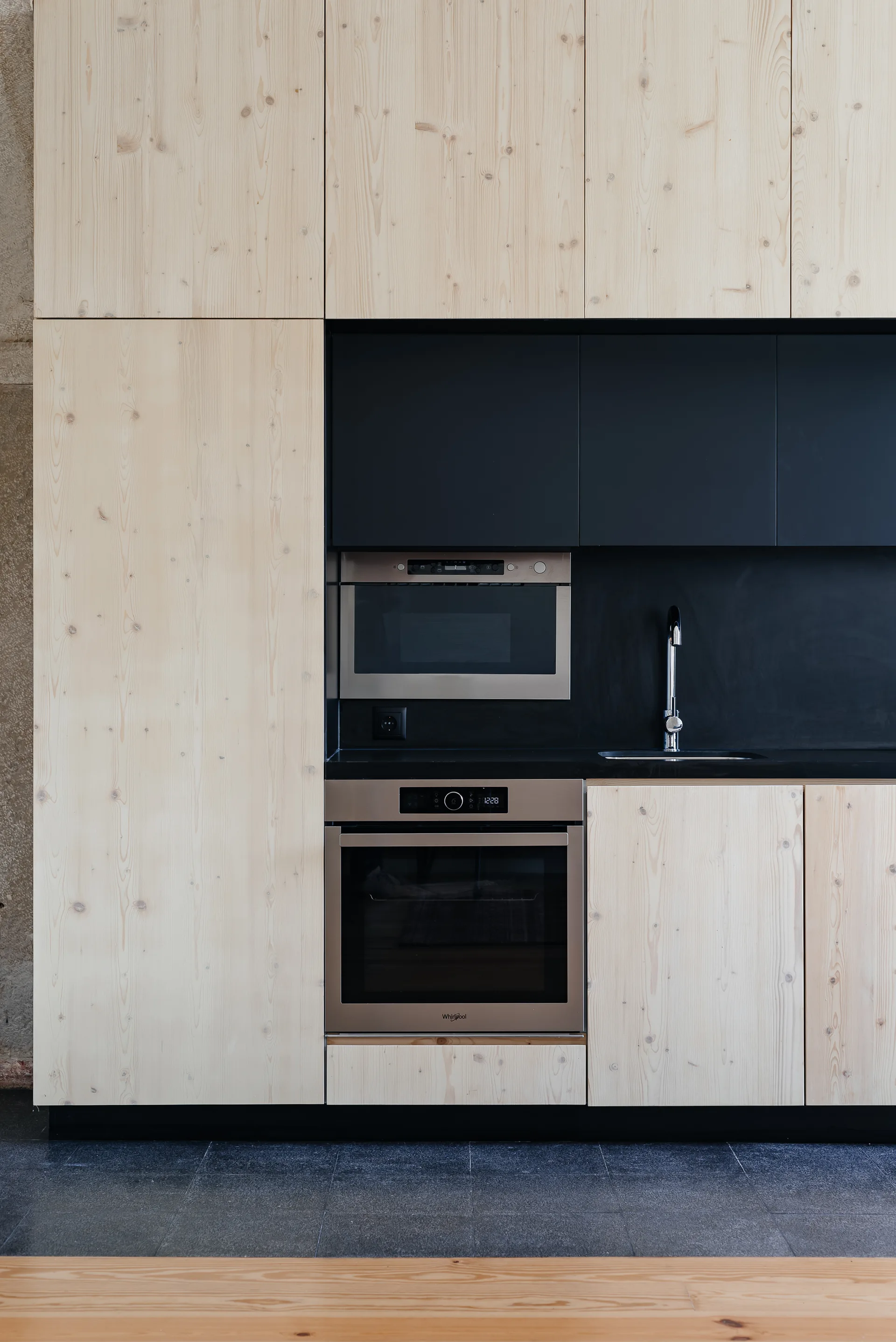 A modern wall of light wood flat-panel kitchen cabinets featuring a matte black inset section with an integrated oven, microwave, and sink.