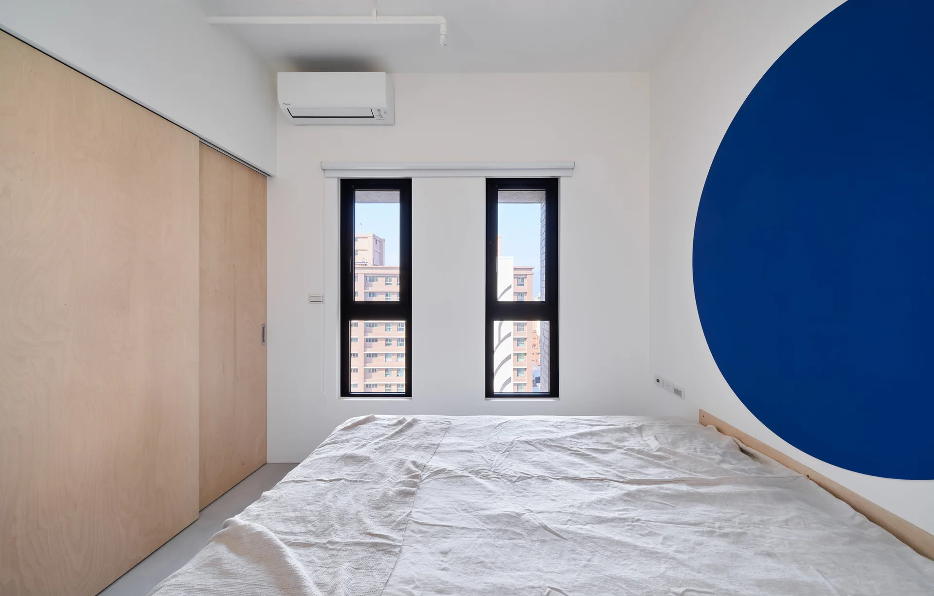 A minimalist bedroom features a bed with white linen bedding, light wood sliding closet doors, and a large blue painted circle on the wall.