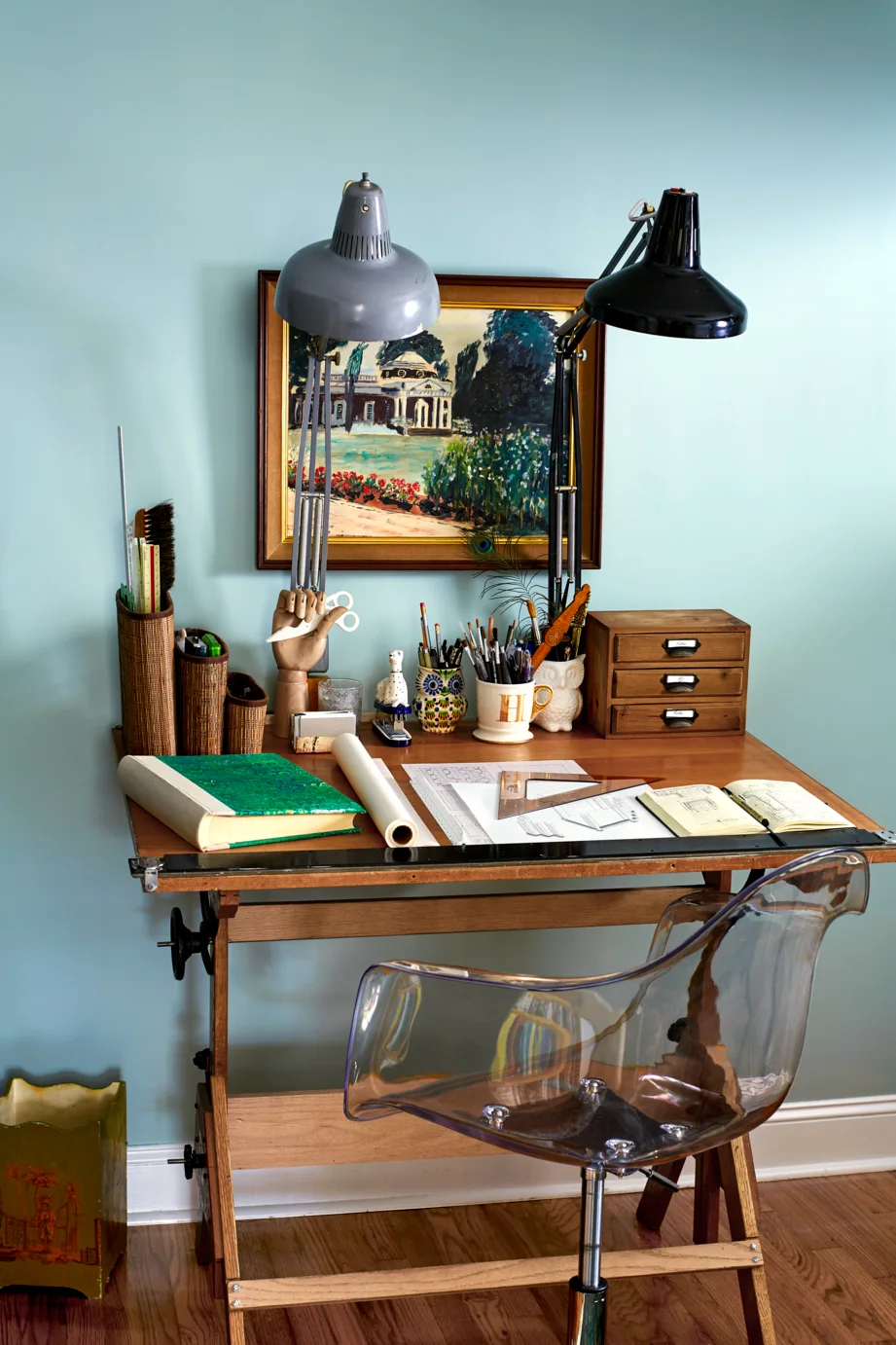 A vintage wood drafting table with black and grey desk lamps, paired with a clear acrylic chair against a light blue wall.