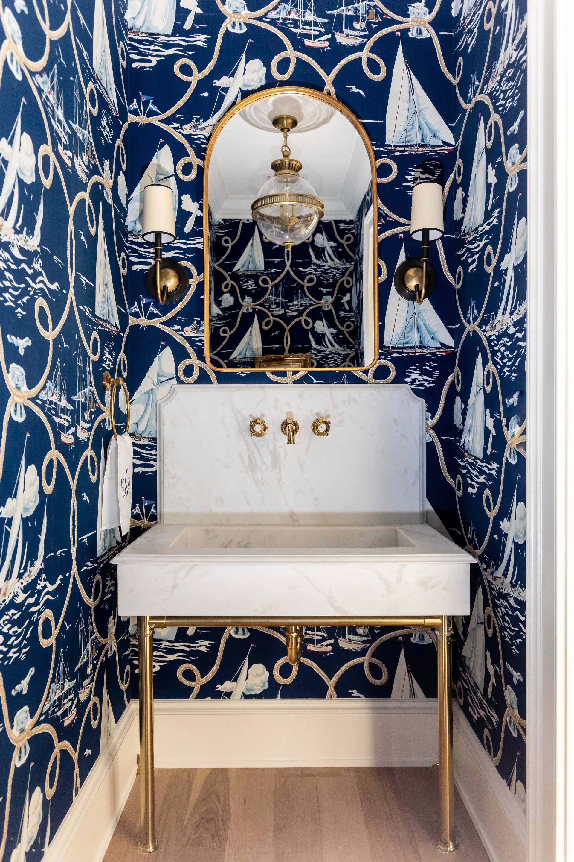 A small powder room features navy blue nautical wallpaper, a marble console sink with brass legs, and an arched brass mirror.