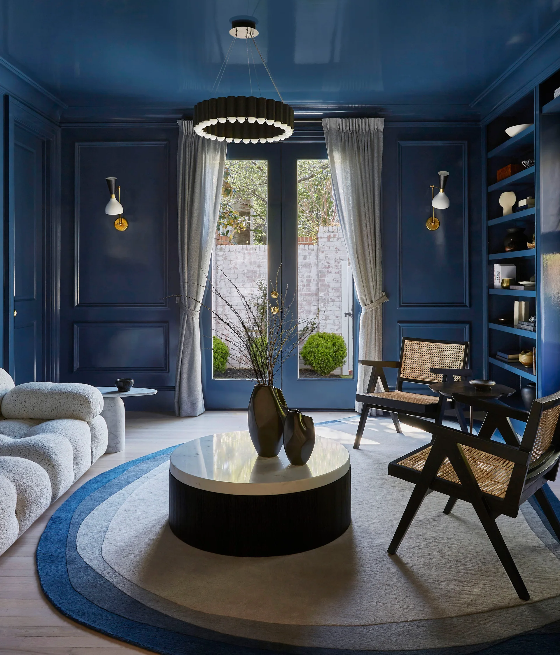 A dark blue living room featuring high-gloss paneled walls, a curved white boucle sofa, cane back armchairs, and a circular gradient rug.