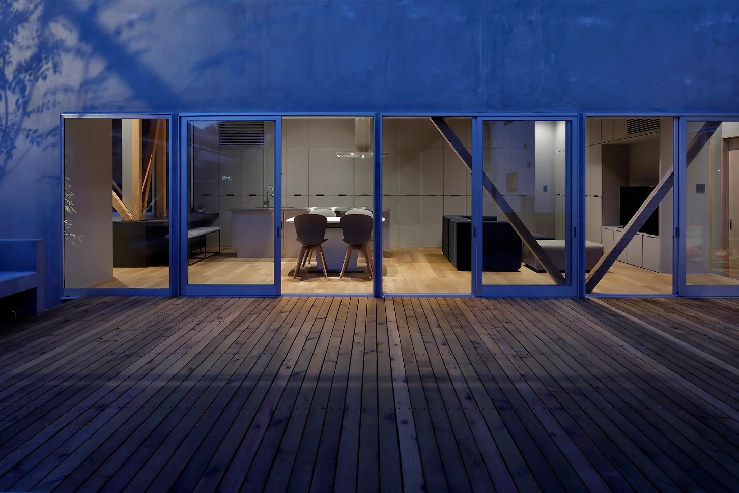 A wide timber deck at twilight features a wall of sliding glass doors looking into a minimalist kitchen and living room.
