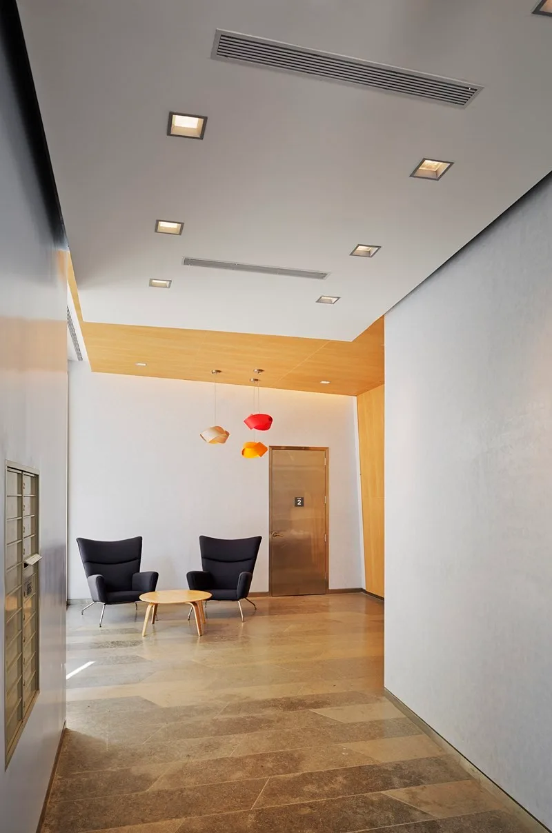 A modern building entry featuring a seating area with two dark gray armchairs, a wooden coffee table, and three colorful pendant lights.