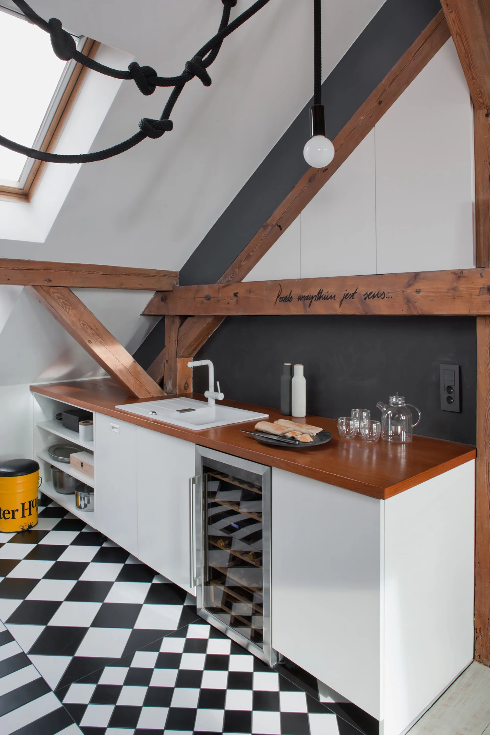 A modern attic kitchen featuring white lower cabinetry, a warm wood countertop, exposed wooden beams, and a black-and-white checkered floor.