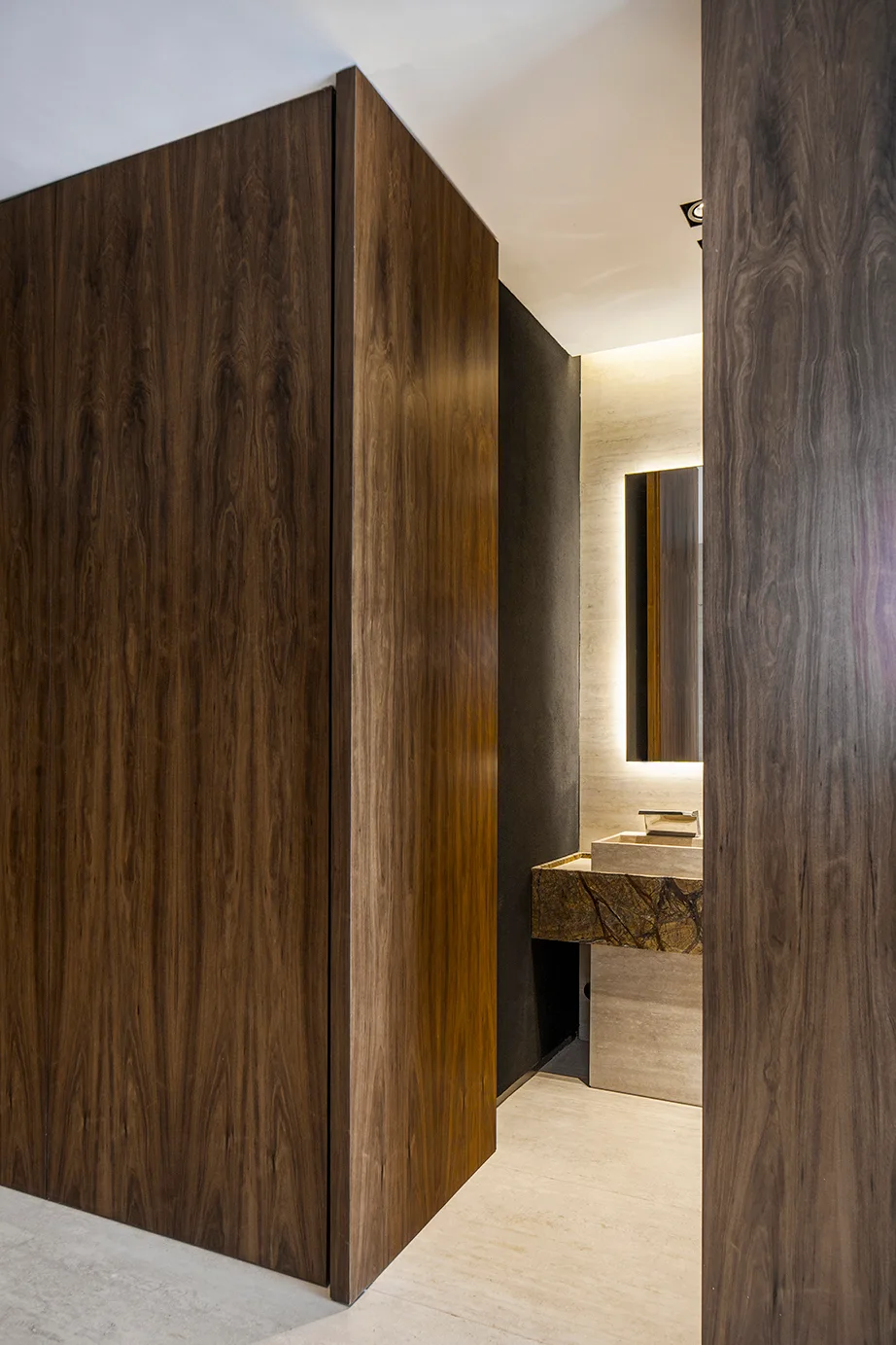 A minimalist powder room entrance featuring tall dark wood paneled walls, a backlit mirror, and a floating stone vanity sink.