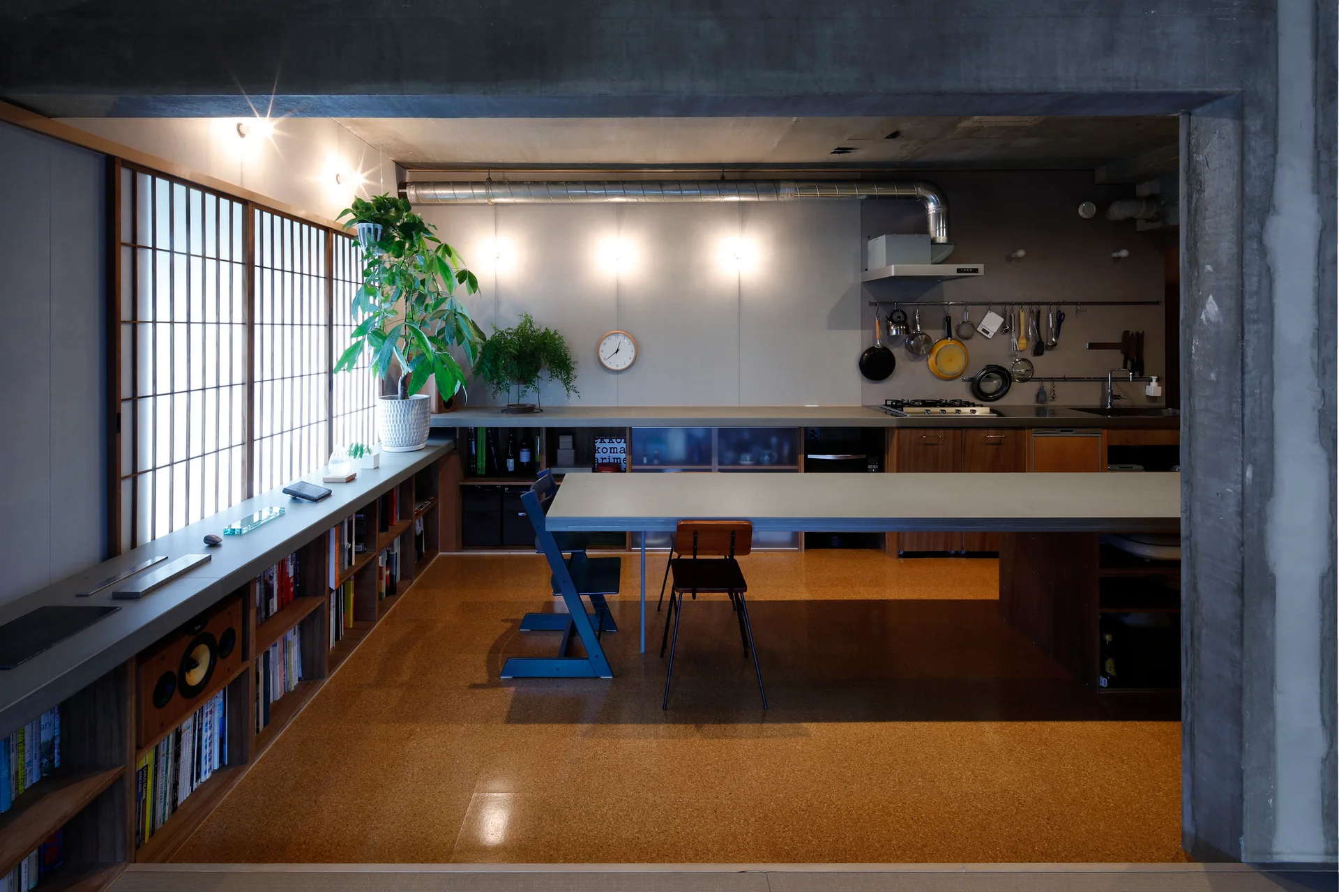 A minimalist kitchen and dining space features warm cork flooring, a long grey counter, built-in bookshelves, and traditional shoji window screens.