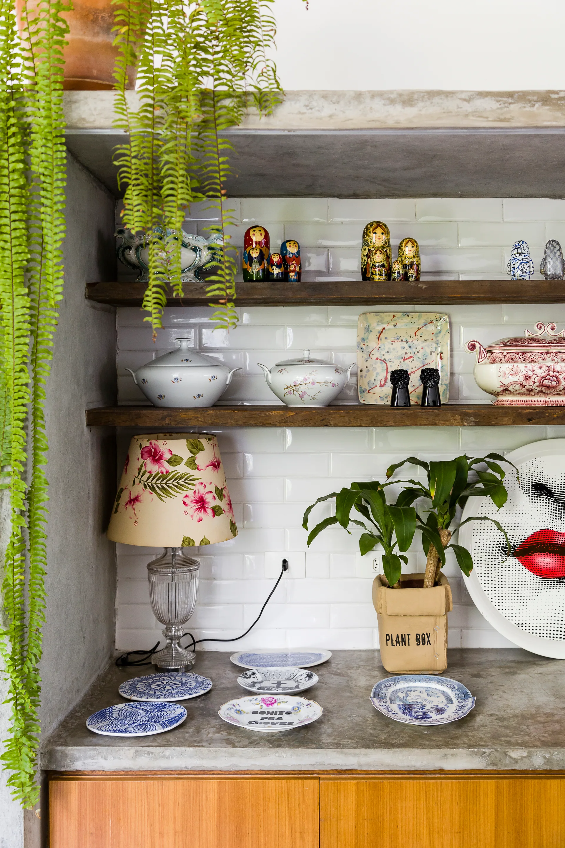 A concrete kitchen counter features wooden shelves against a white subway tile backsplash decorated with ceramic dishes, a floral lamp, and plants.