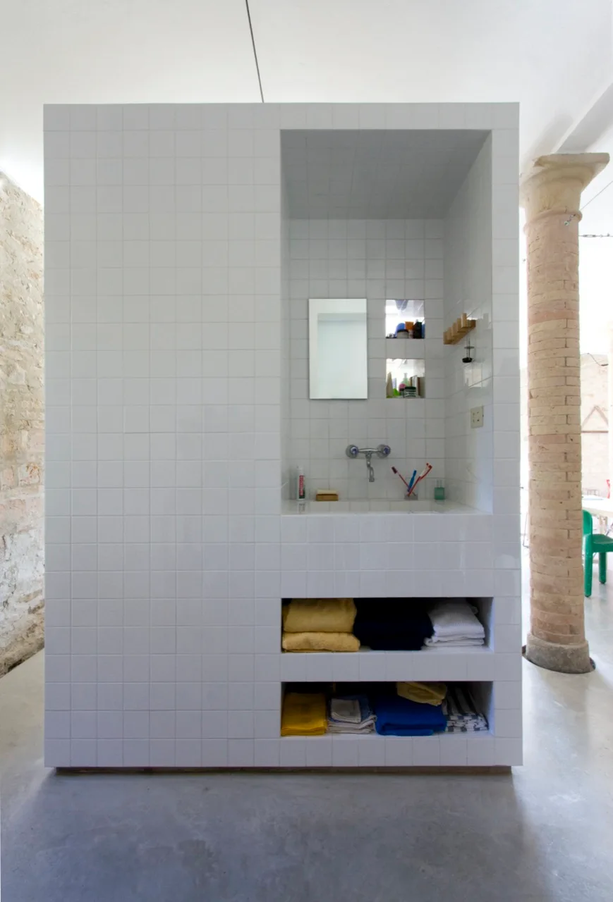 A freestanding white tiled vanity unit with an integrated sink, rectangular mirror, and open storage shelves stands on a concrete floor.