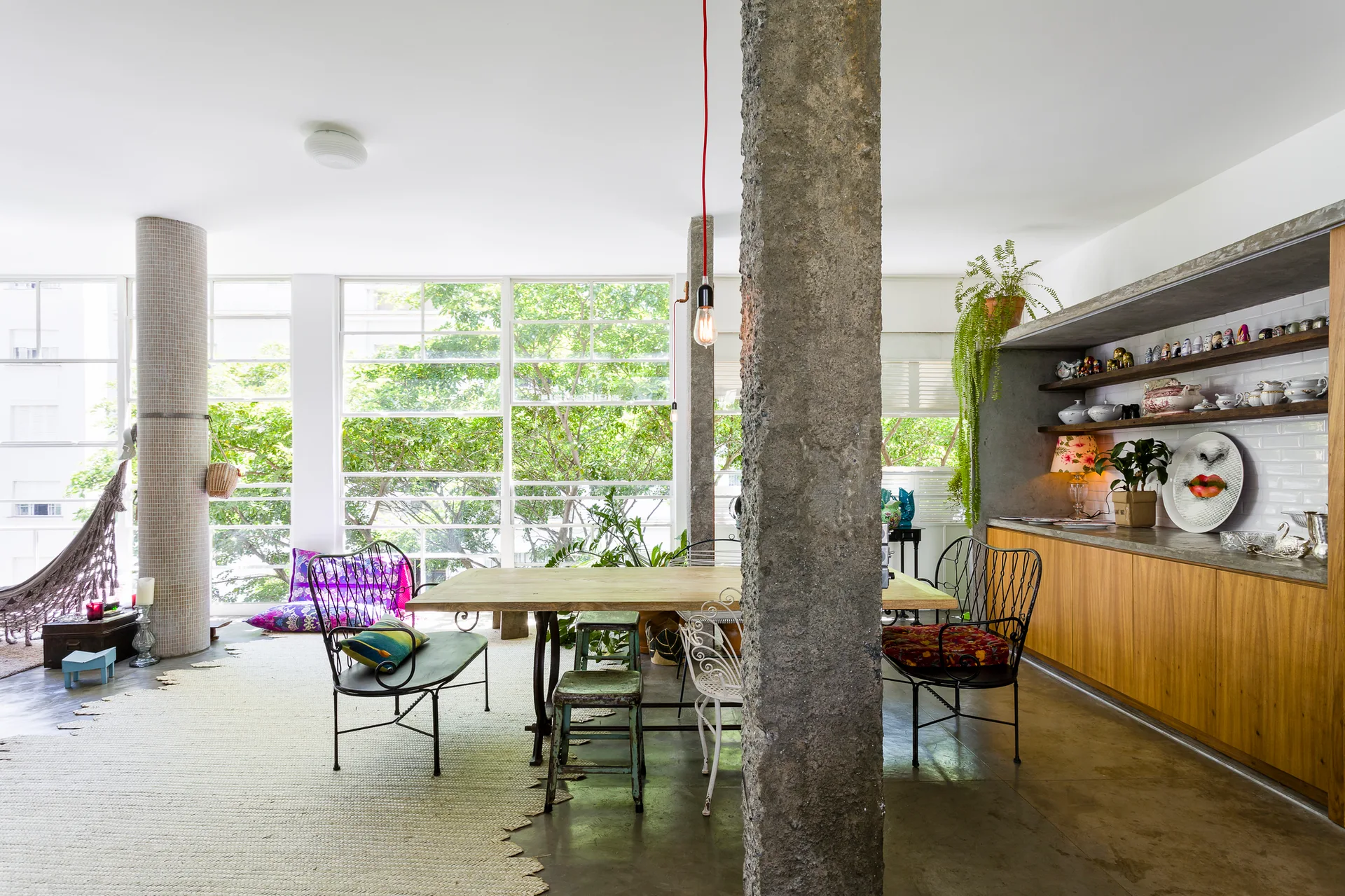 An eclectic dining area features a wooden table with mismatched chairs, exposed concrete columns, and large windows overlooking green trees.