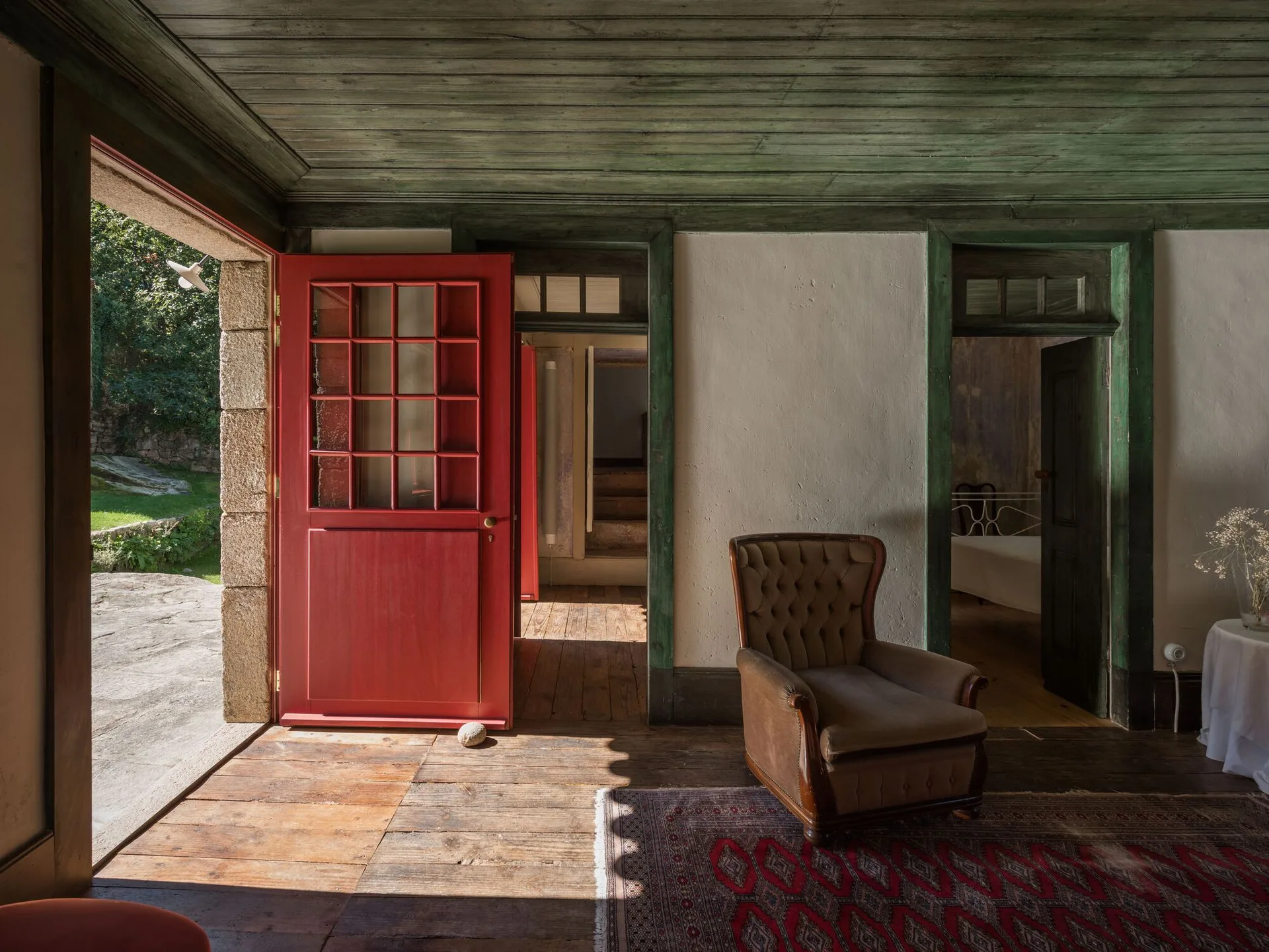 A rustic living room featuring a green painted wood plank ceiling, an open red door, a tufted armchair, and an oriental rug on wood floors.
