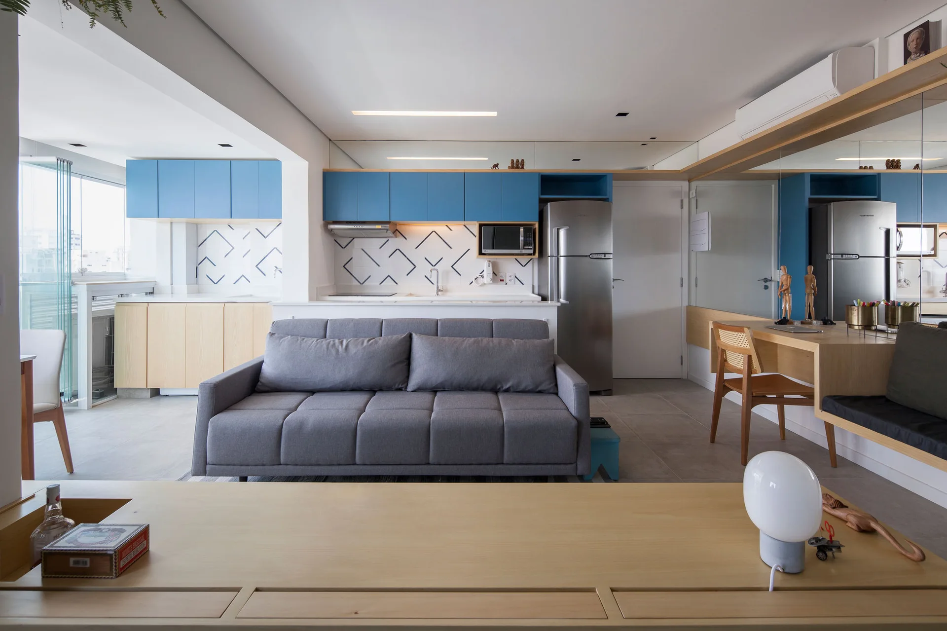 An open-concept living area with a grey sofa, light wood desk, a large wall mirror, and blue kitchen cabinets with a geometric tiled backsplash.