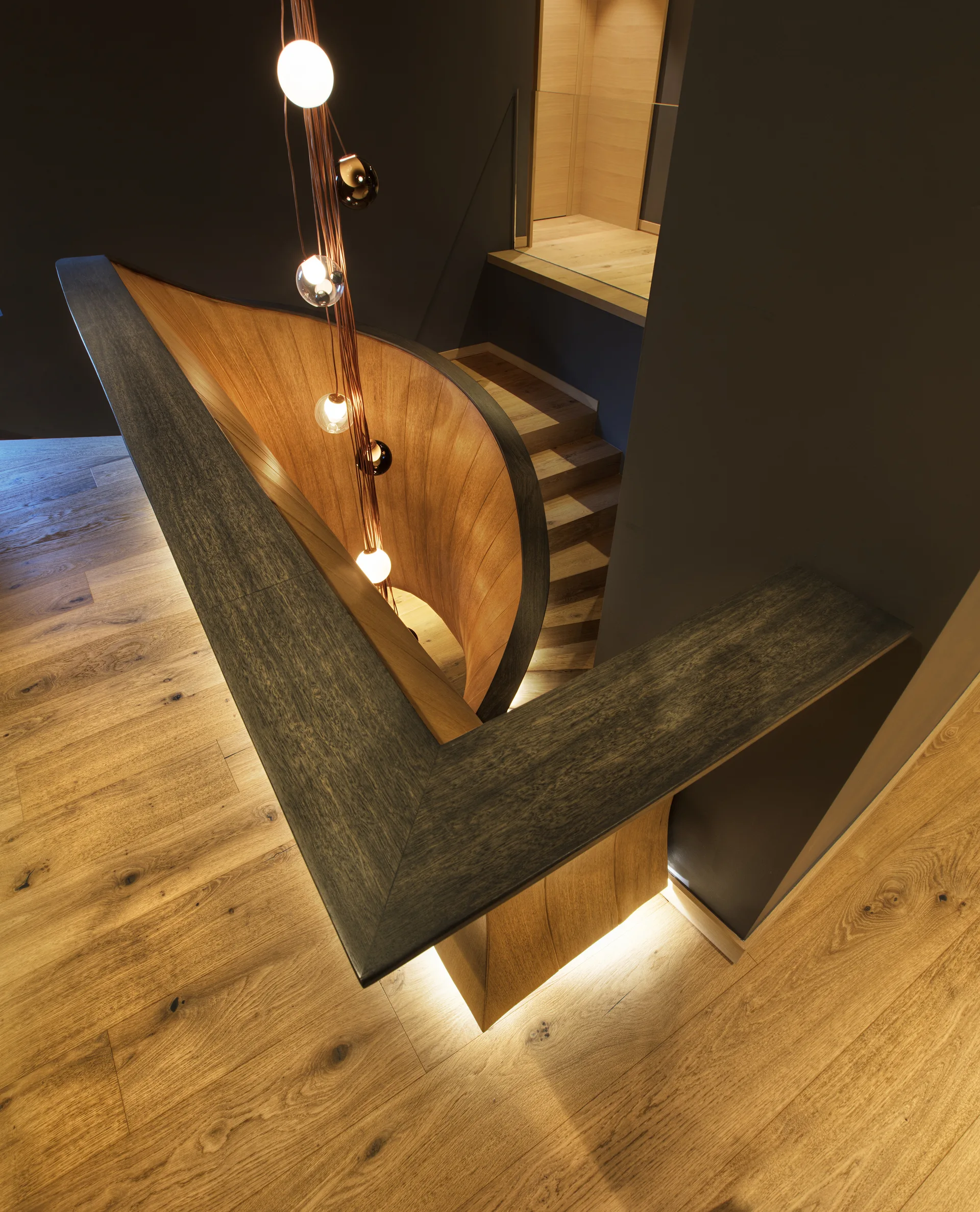 A high-angle view of a curved wooden staircase with a cascading orb pendant light, dark gray walls, and illuminated baseboard lighting.