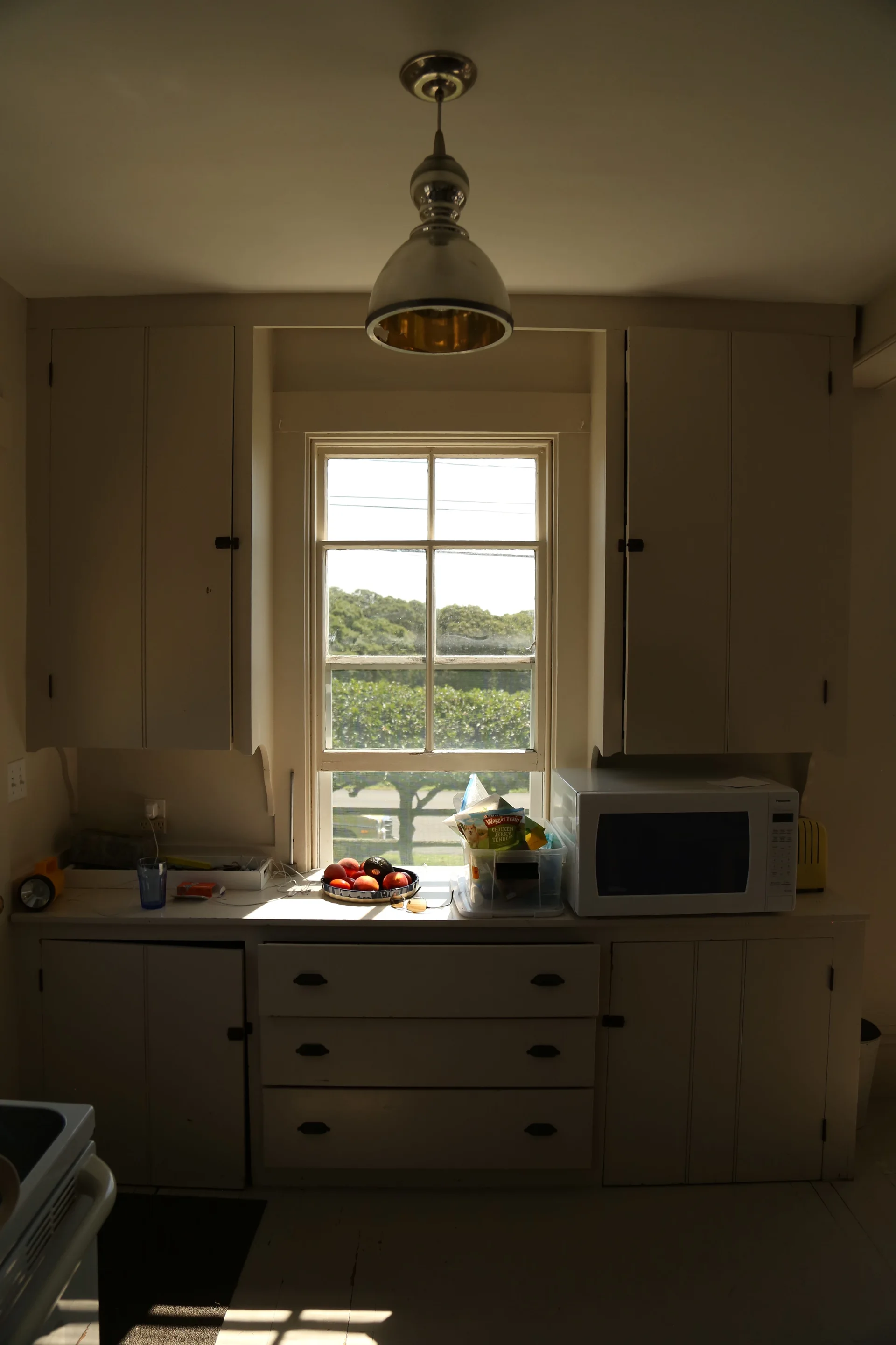 Symmetrical off-white kitchen cabinetry flanks a central multi-pane window with a dome pendant light overhead and a microwave on the counter.