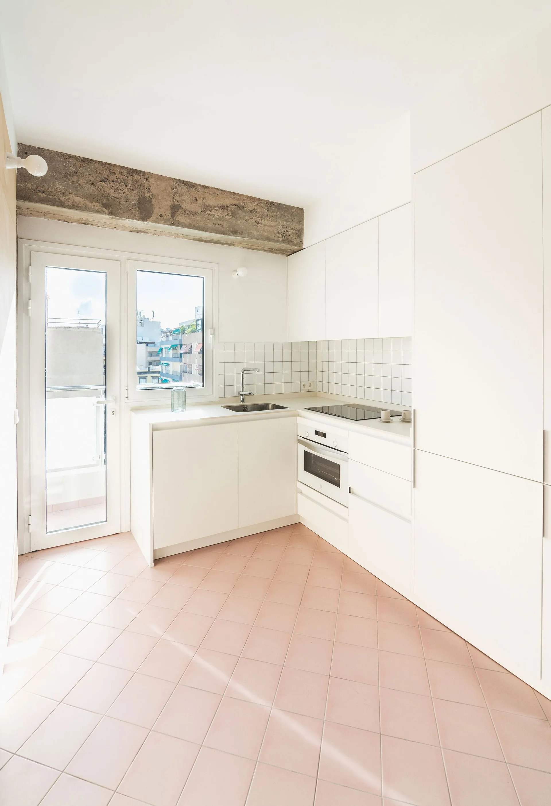 A minimalist white kitchen featuring an exposed concrete ceiling beam, flat-front cabinets, and a pink tile floor.