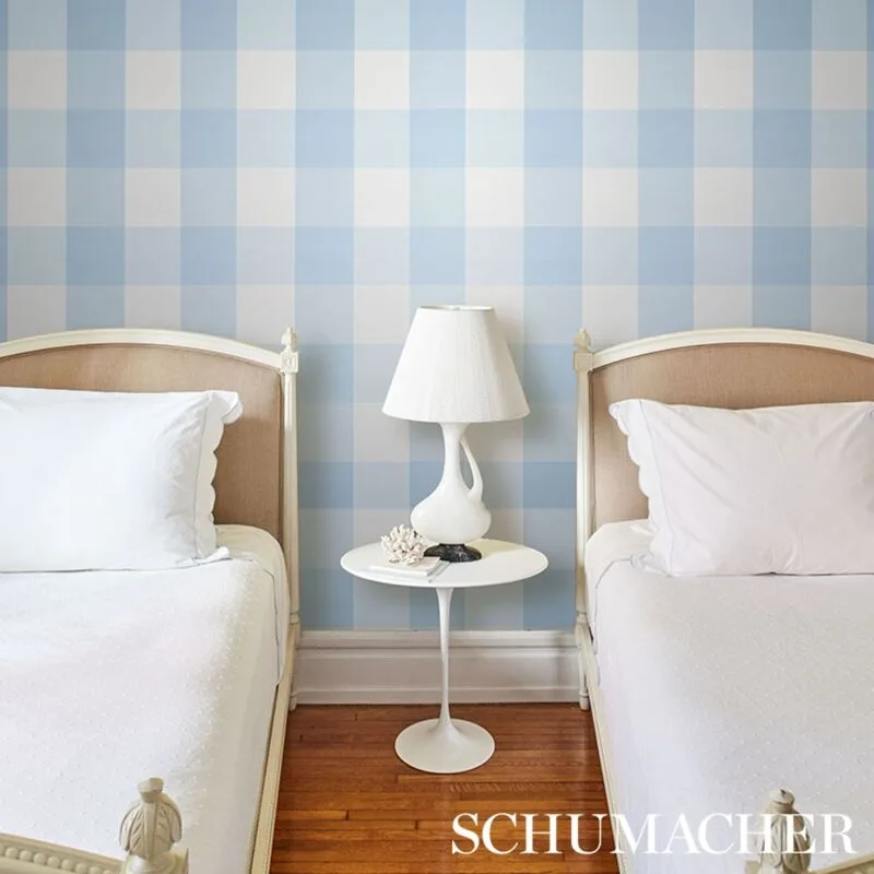 Two twin beds with tan upholstered headboards flank a white pedestal side table in front of a blue and white checkered wallpapered wall.