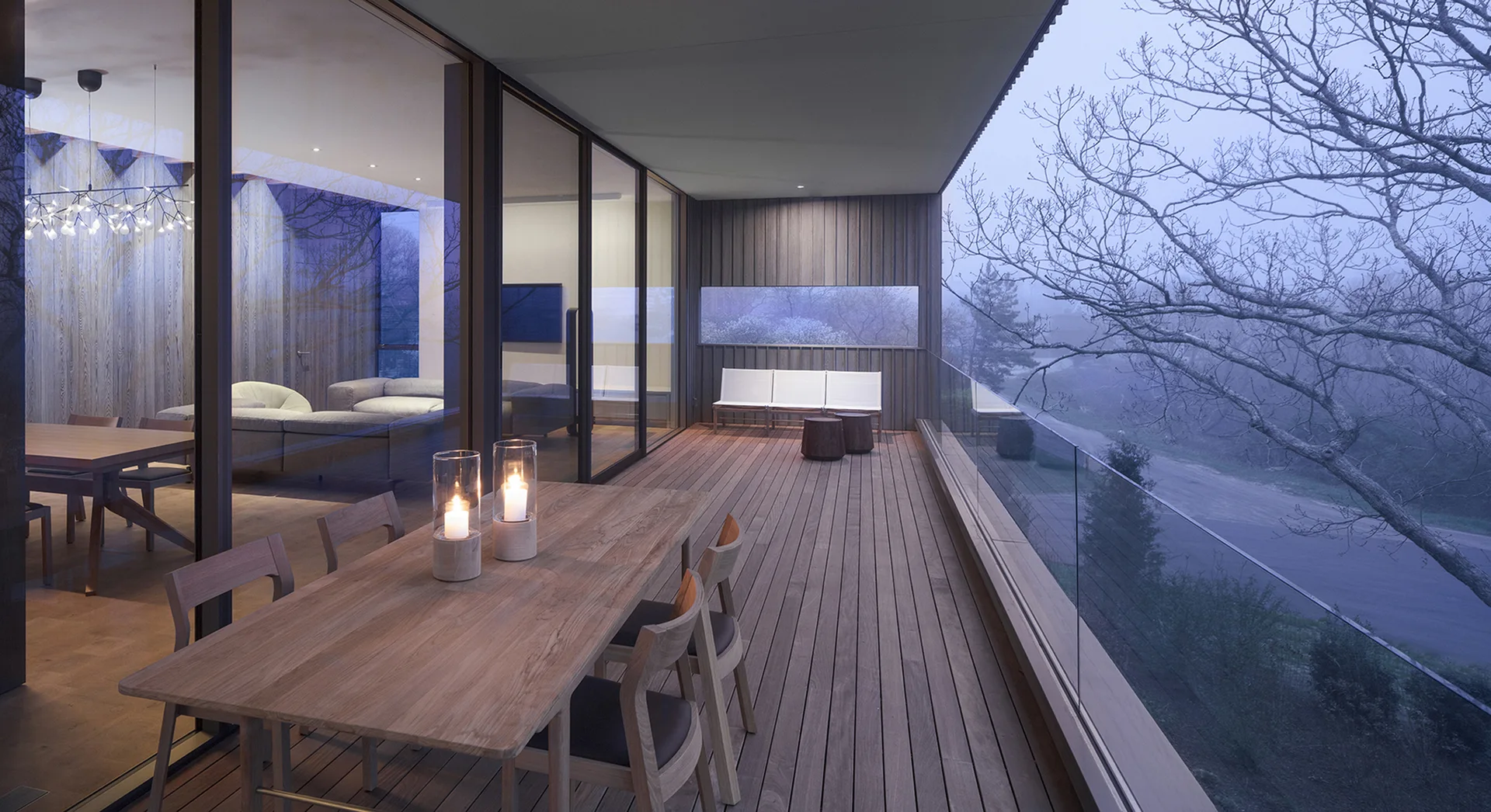 A wooden terrace features an outdoor wood dining table with candles, glass sliding doors, and a glass railing overlooking misty trees.
