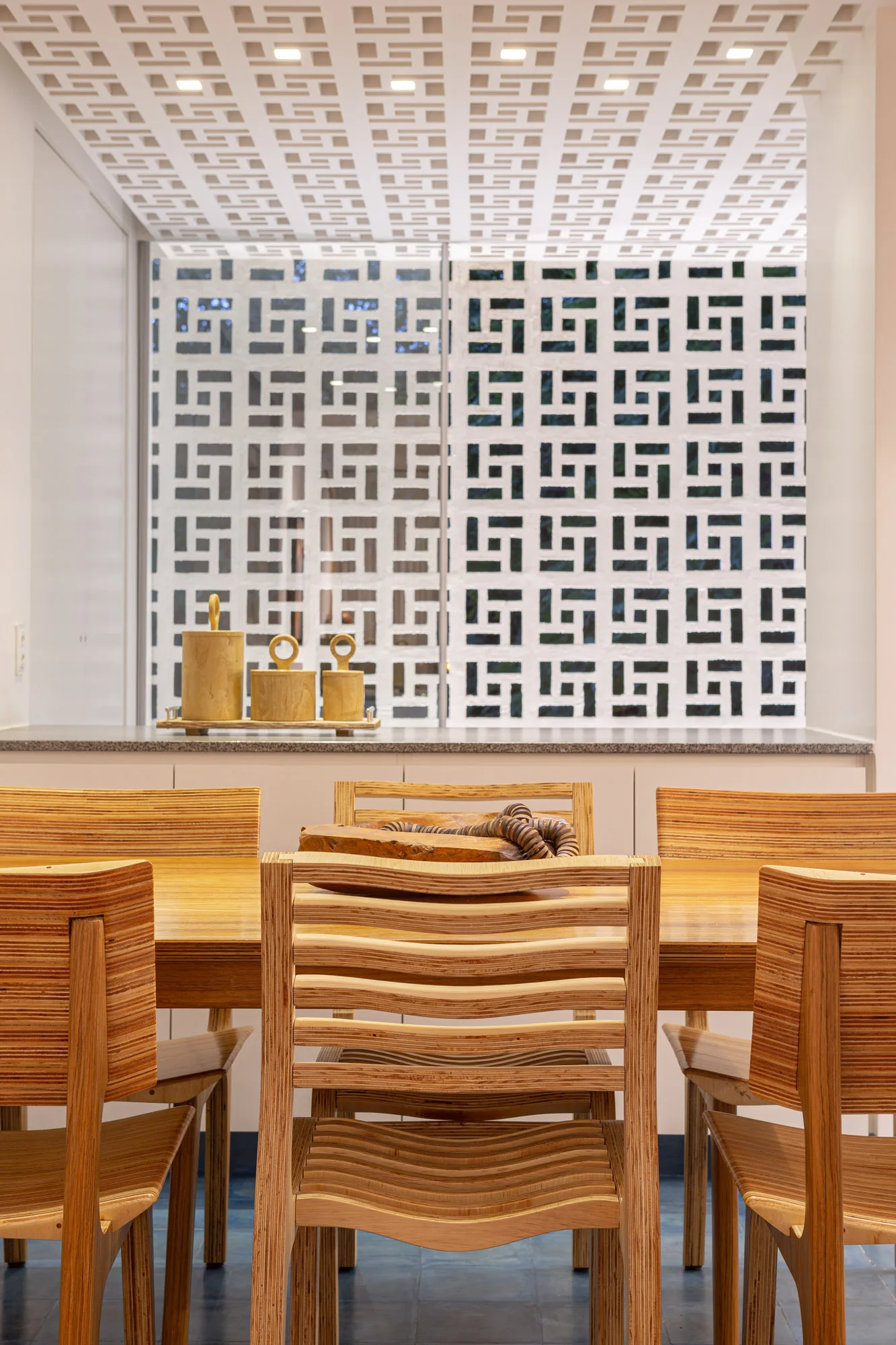 Layered plywood chairs surround a wooden dining table in front of a white geometric breeze block screen wall.
