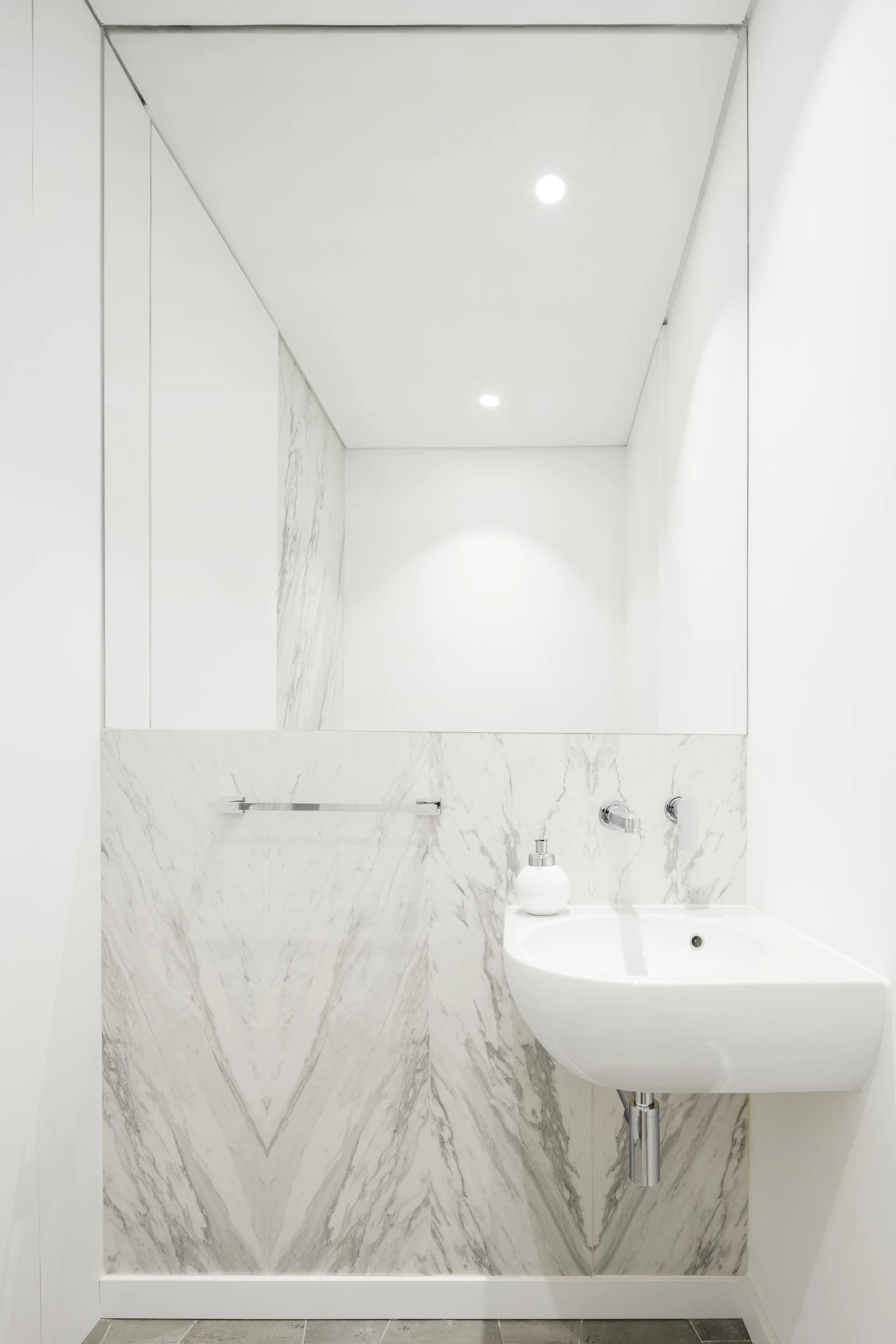 A wall-mounted white ceramic sink is set against grey-veined marble wall cladding beneath a large frameless mirror in a minimalist bathroom.
