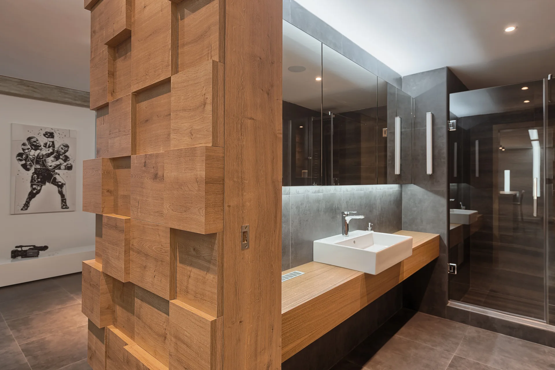 A modern bathroom features a floating wood vanity with a white vessel sink next to a textured 3D wood block wall divider and grey tile.