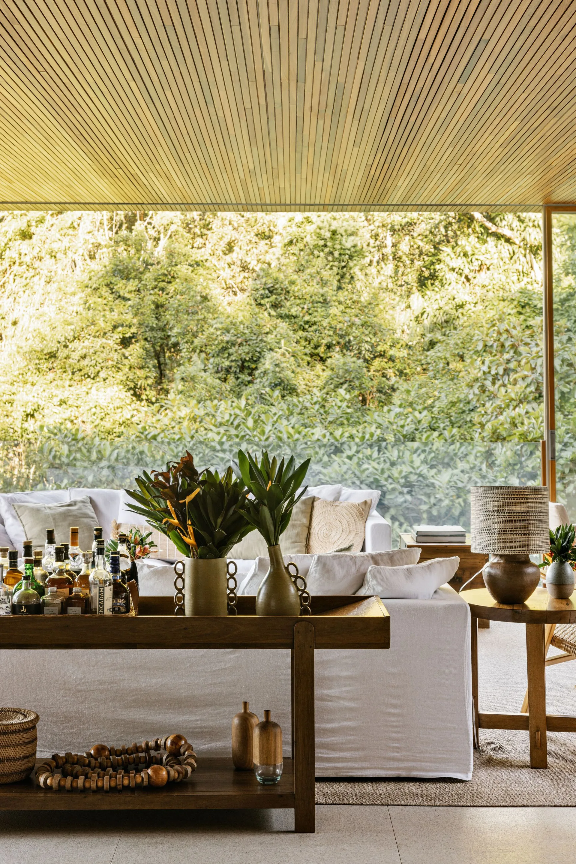 A bright living space features a white sofa, a wooden console table with bottles, and a slatted wood ceiling overlooking green outdoor plants.