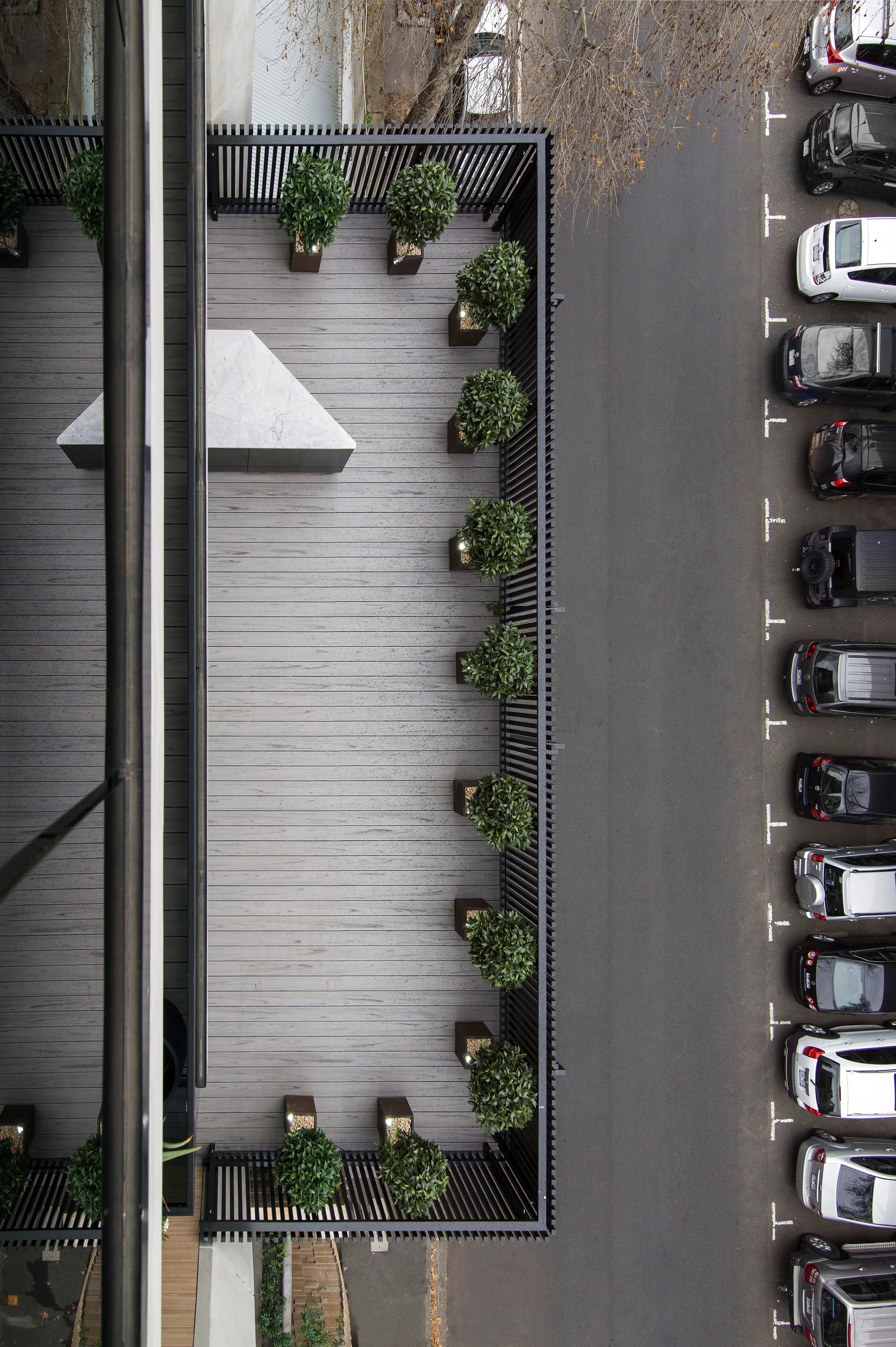 Overhead view of a minimalist grey terrace deck with a row of potted green shrubs and black metal slat fencing.