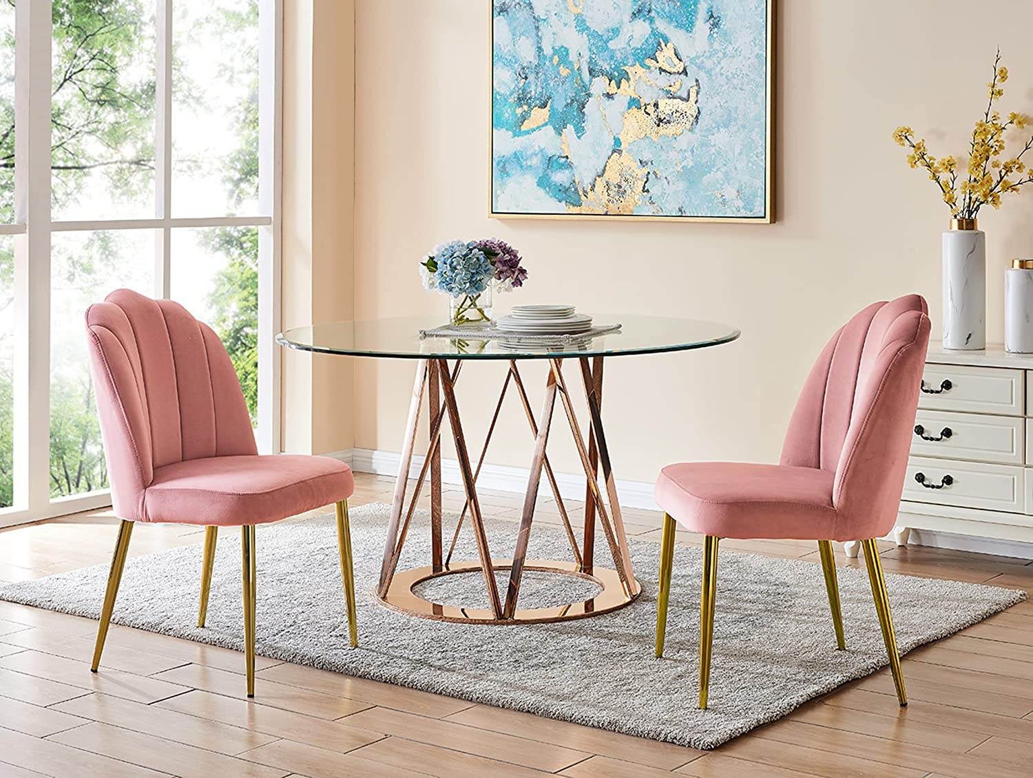 A round glass-top dining table with a gold metal base sits between two blush pink velvet chairs on a grey textured rug.