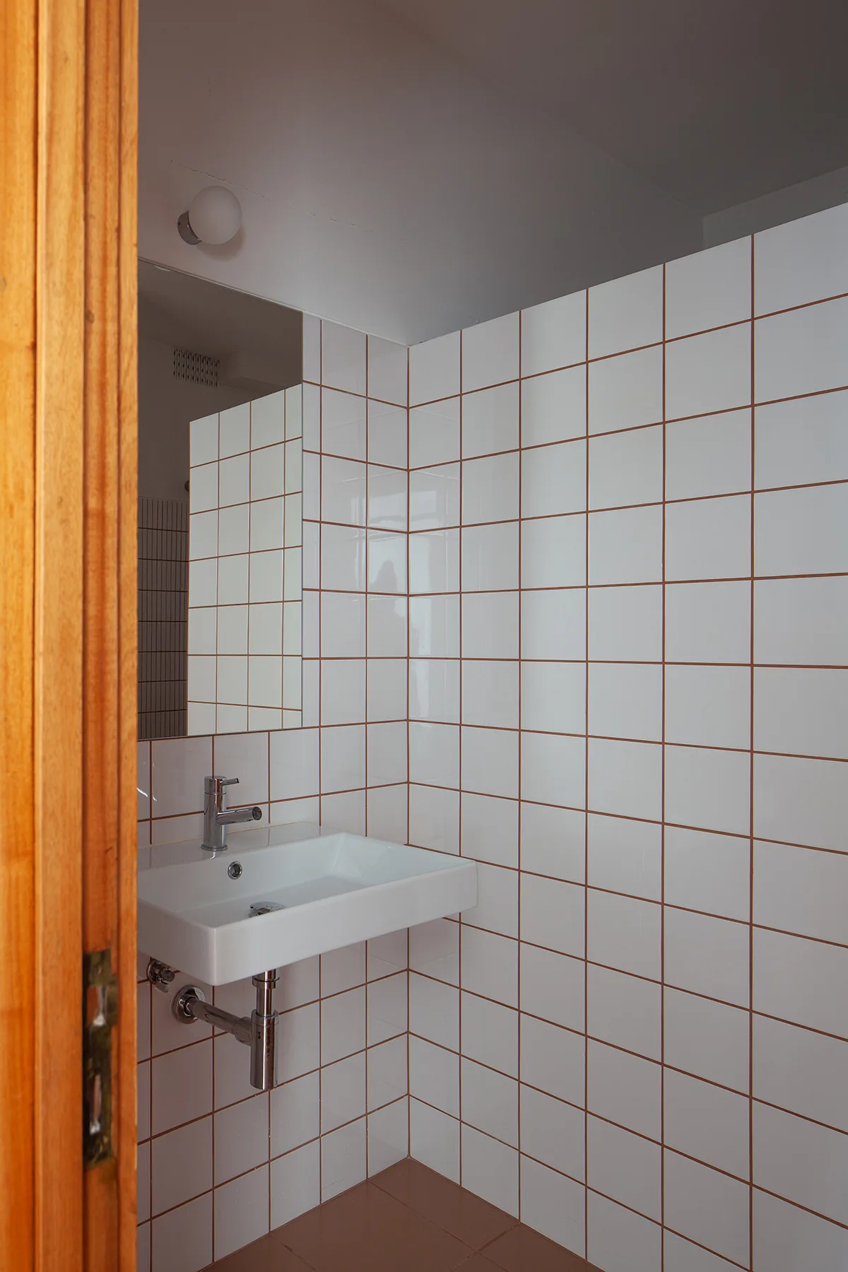 A minimalist bathroom featuring white square grid wall tiles, a wall-mounted ceramic sink with a chrome faucet, and a large mirror.