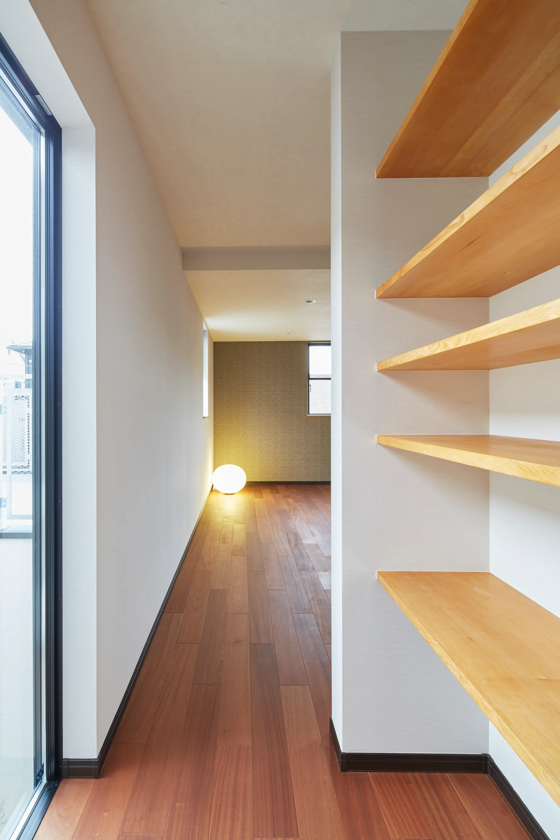 A minimalist hallway features dark wood flooring, open built-in wooden shelves, and a spherical floor lamp glowing at the far end.