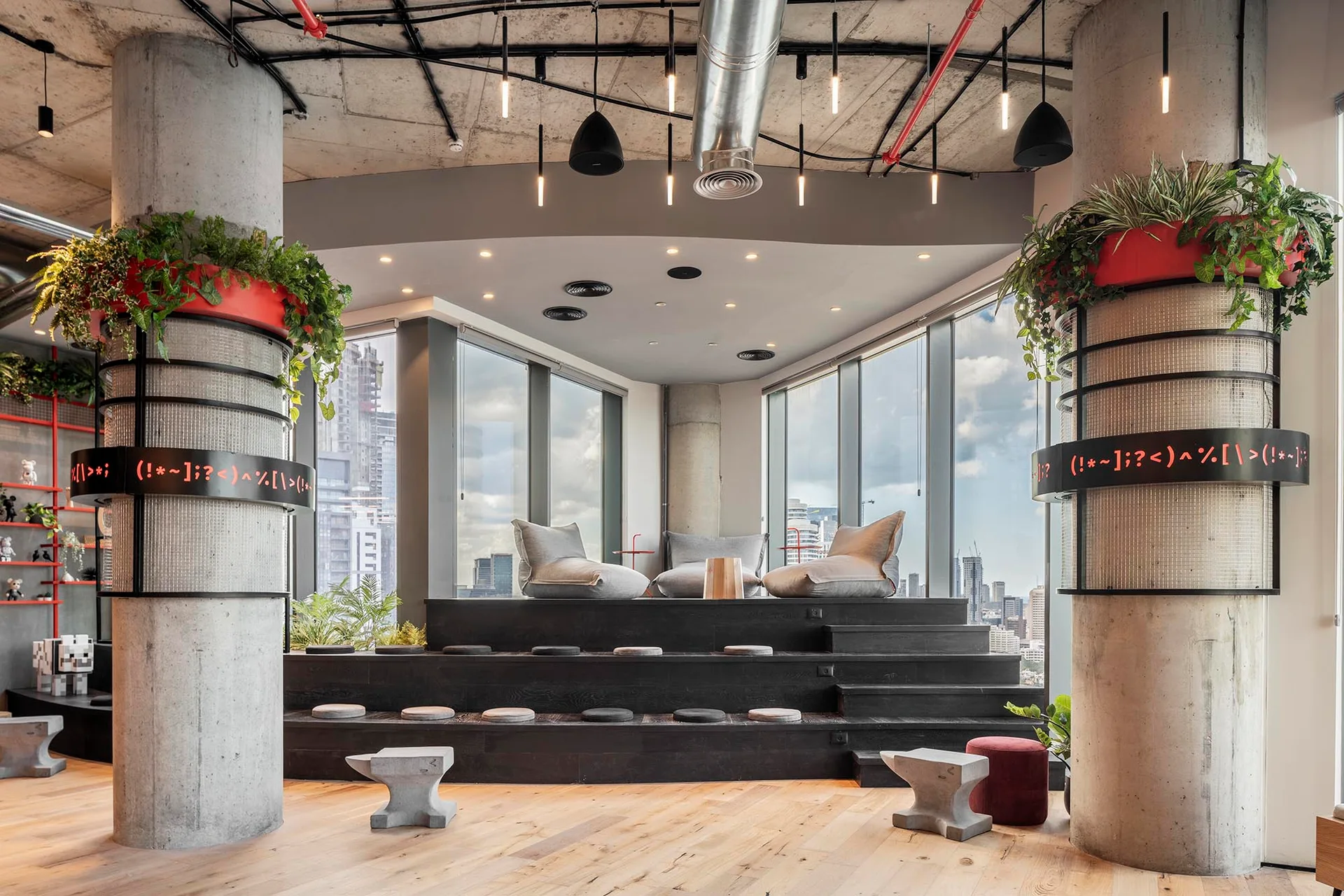 A modern lounge features a black tiered wooden seating platform with bean bags, flanked by large concrete columns with red planters under an exposed ceiling.